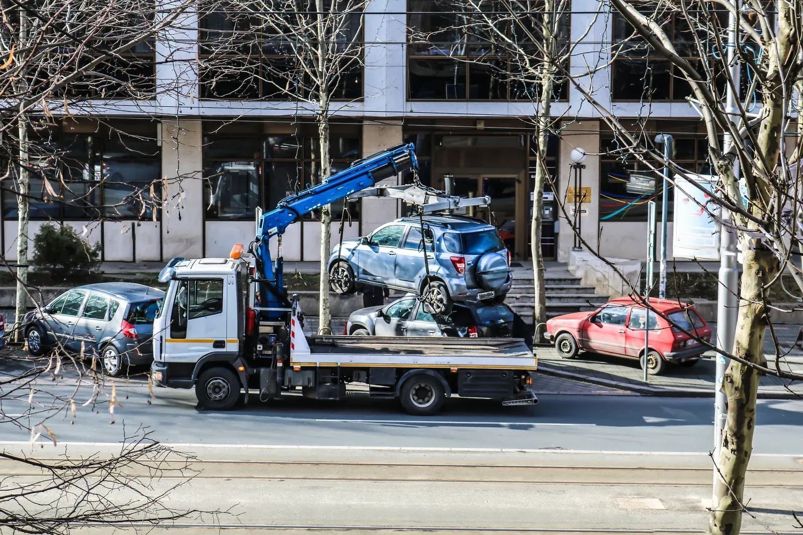 Tow truck lifting a blue SUV. Several other cars are parked on the street in front of a building.