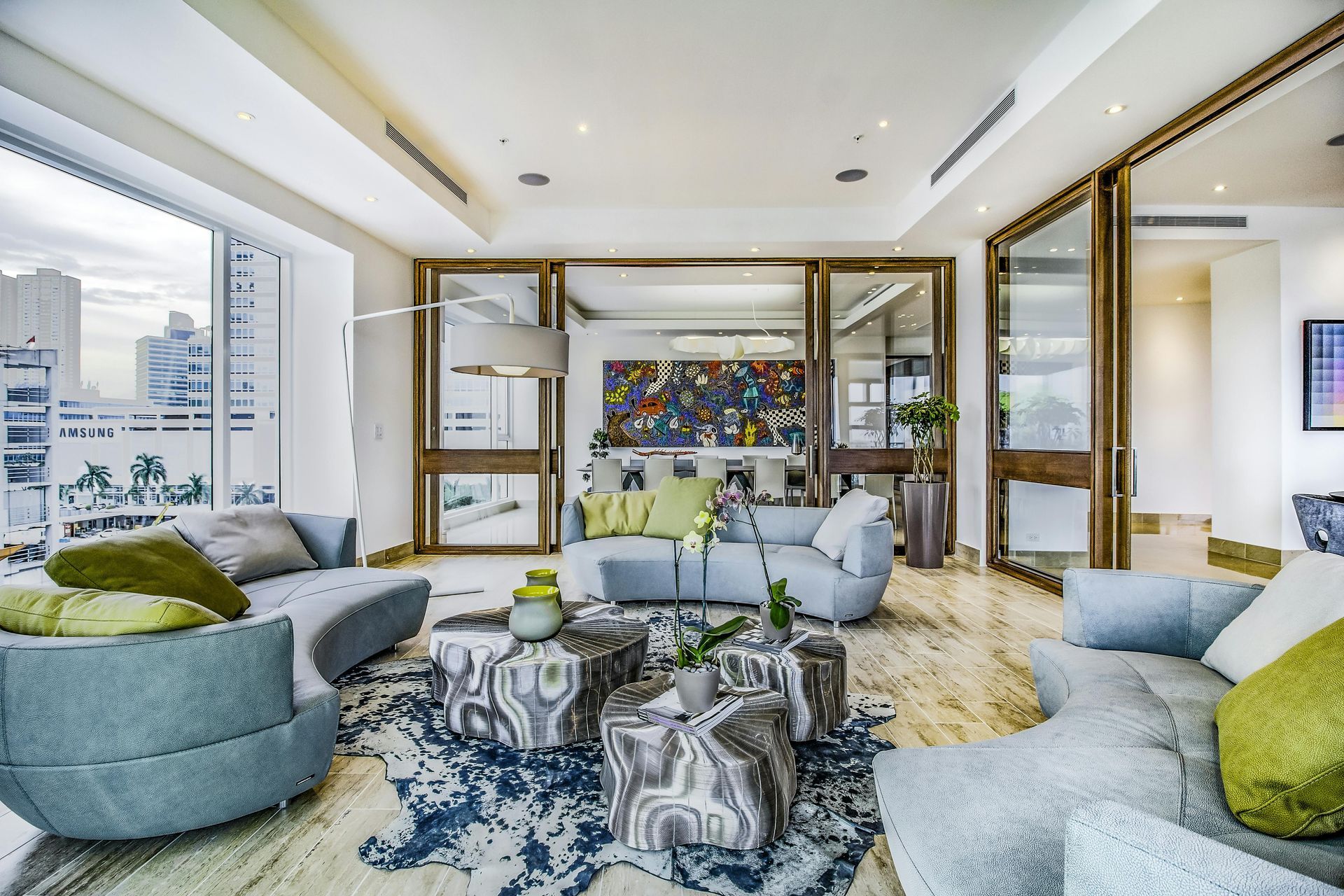Spacious, modern living room with blue and green sofas, patterned rug, and large glass doors opening to a city view.