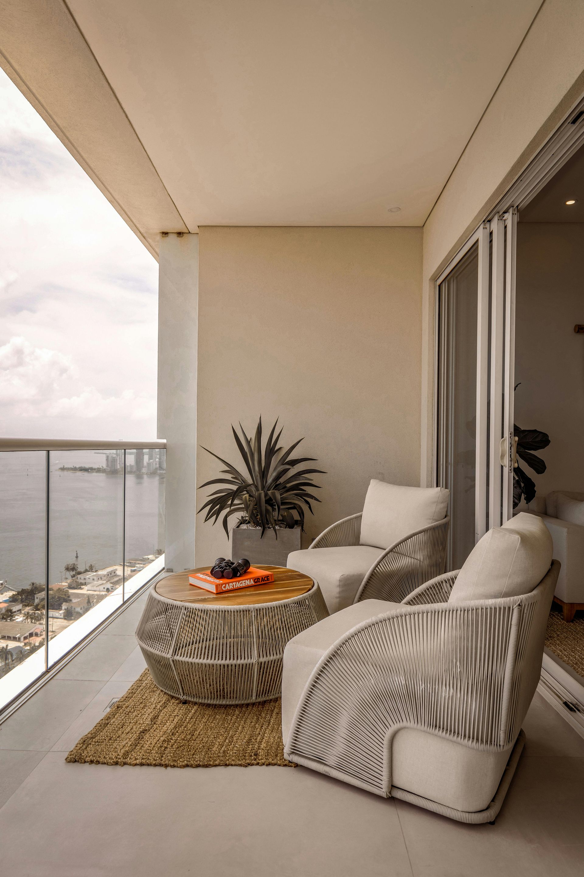Balcony with two woven armchairs, round coffee table, and a potted plant. Overlooks a waterfront view with a glass railing.