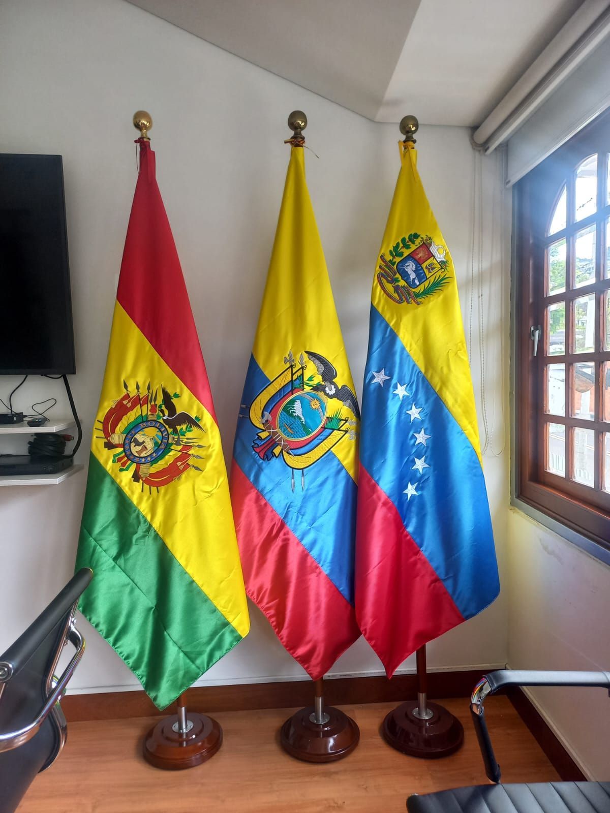 Tres banderas de pie en una habitación: Bolivia, Ecuador y Venezuela, junto a una ventana.