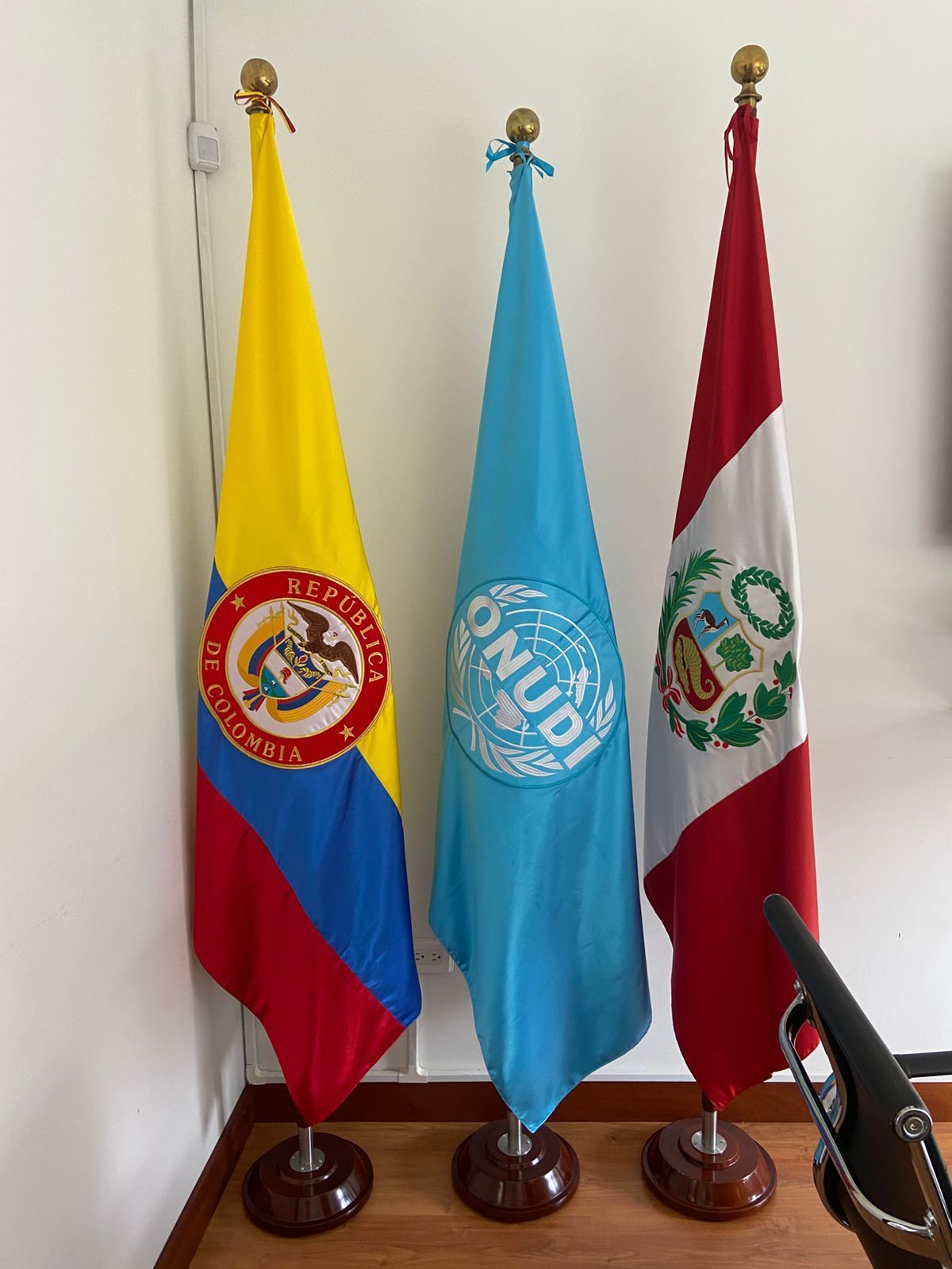 Tres banderas sobre soportes en un pasillo: Colombia, ONU y Perú.