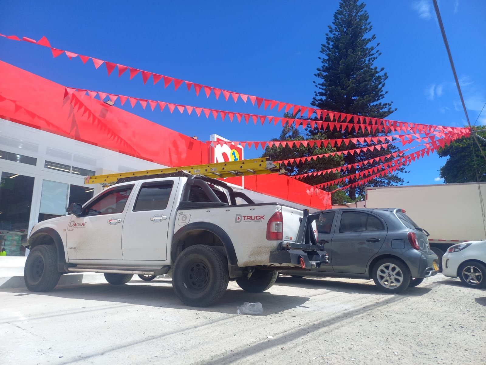 Camioneta blanca y SUV gris estacionadas bajo banderines rojos en un lote soleado.