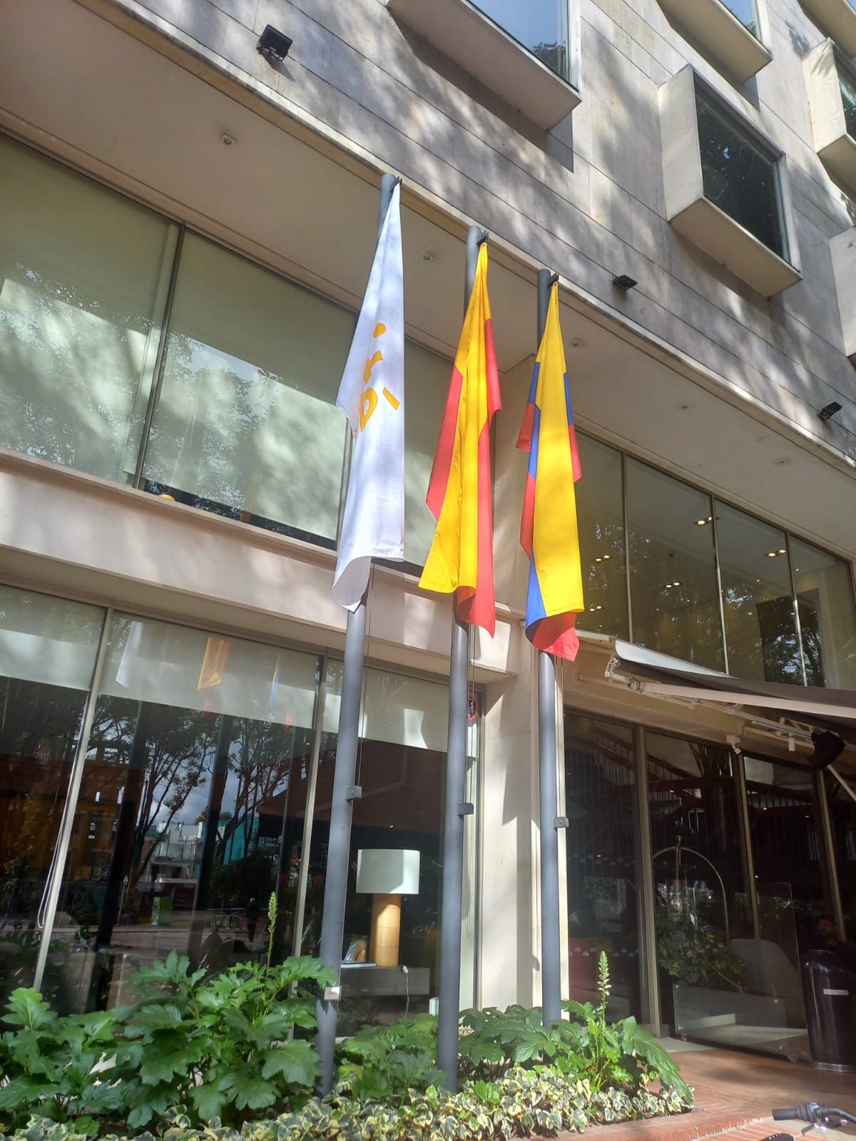 Tres banderas junto a la entrada de un edificio con puertas y ventanas de cristal, bordeado de vegetación.