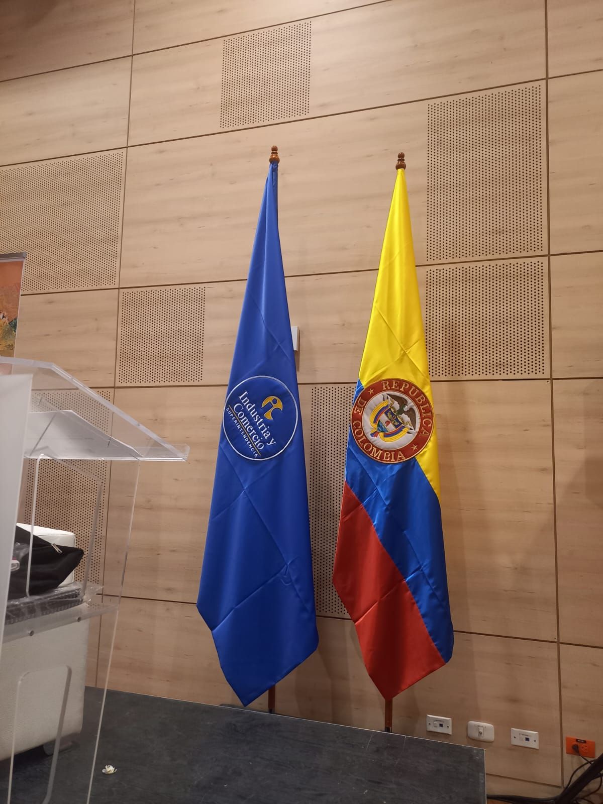 Dos banderas sobre soportes junto a un atril transparente en una sala de conferencias con paneles de madera.