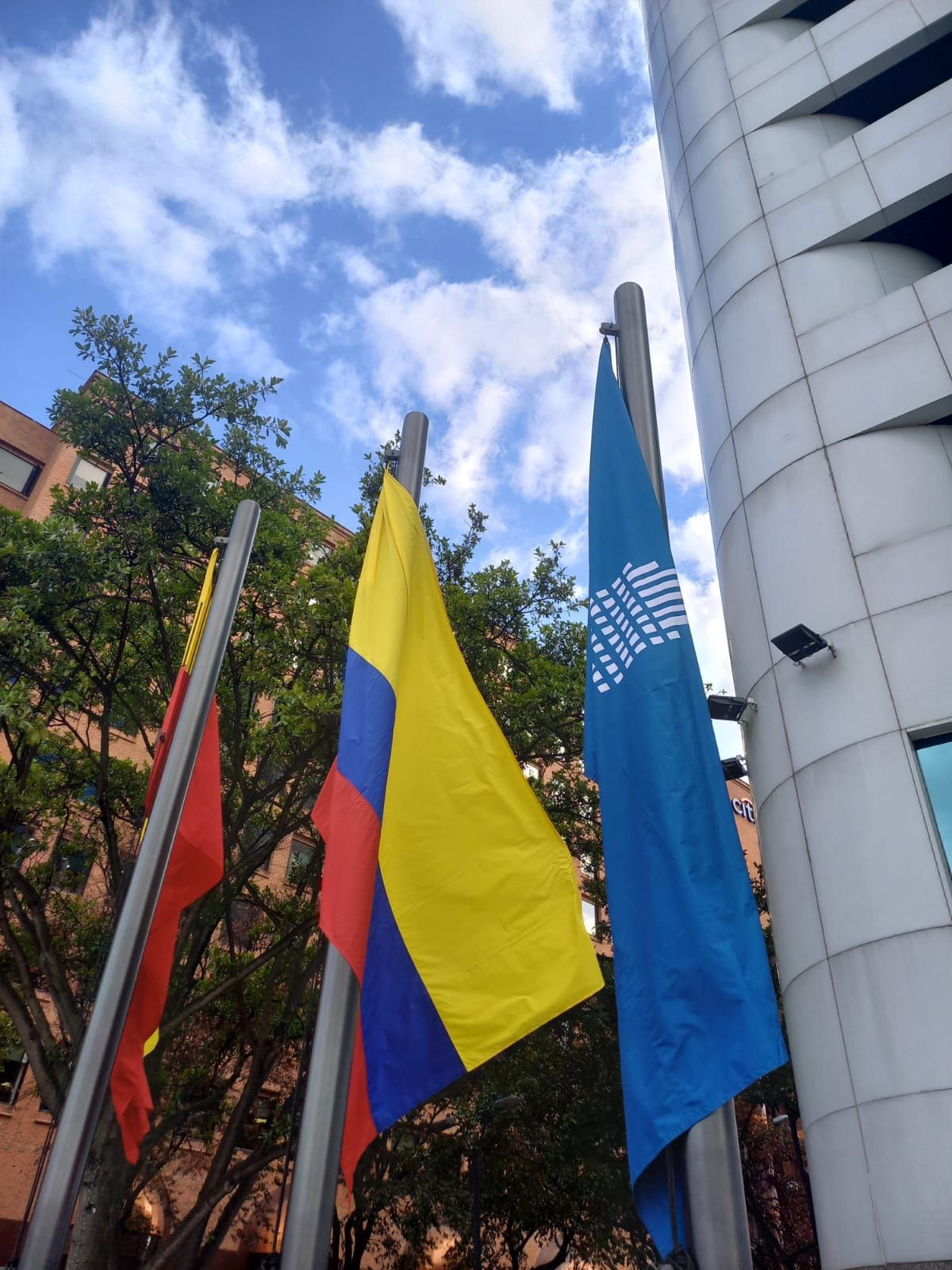Tres banderas, incluida la bandera amarilla, azul y roja de Colombia, frente a un edificio moderno bajo un cielo azul.