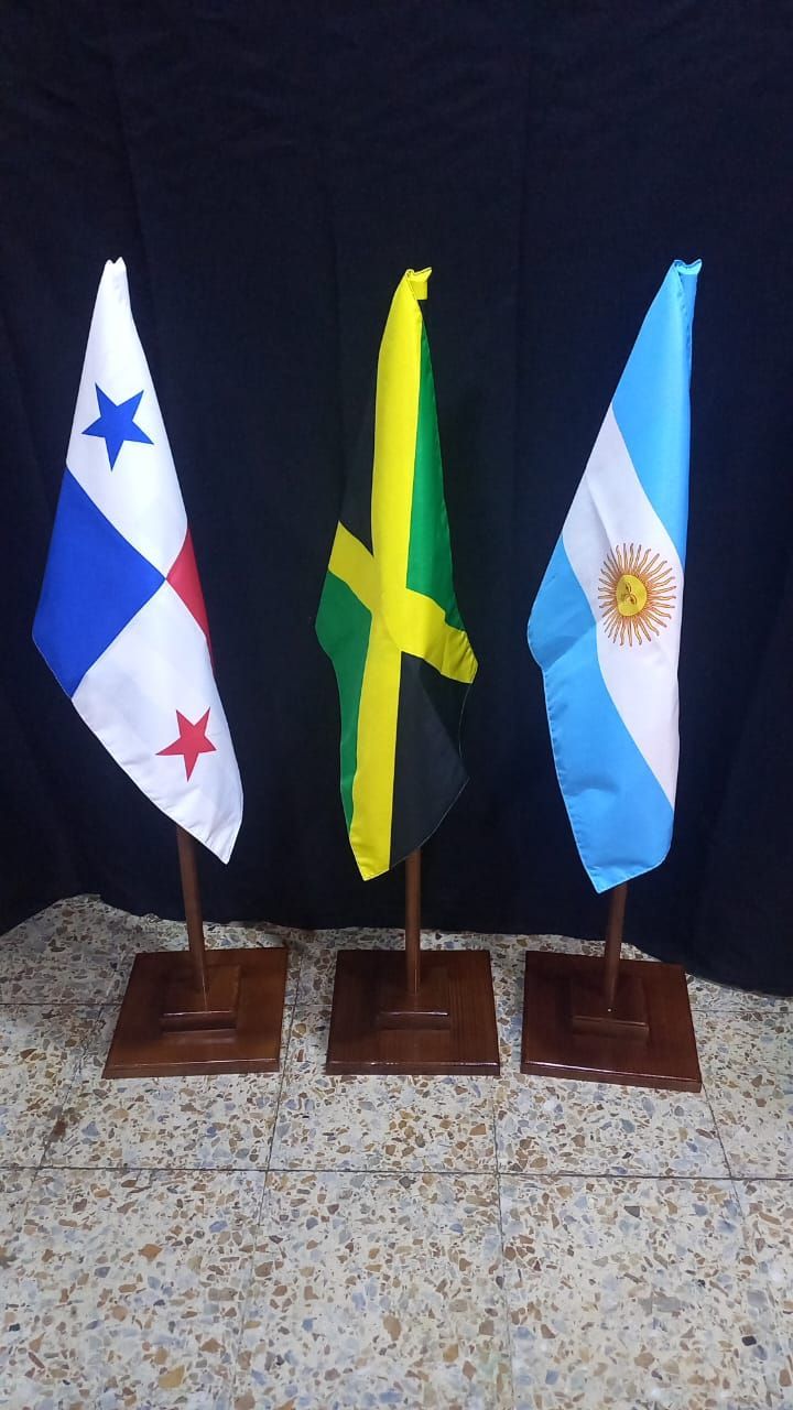 Tres pequeñas banderas de mesa sobre bases de madera: Panamá, Jamaica y Argentina, sobre un fondo oscuro.