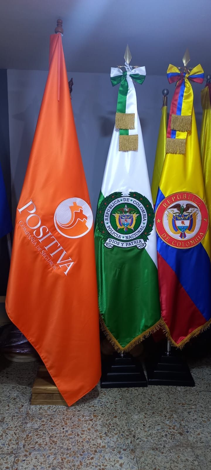 Tres banderas de pie en naranja, verde y amarillo-púrpura-rojo junto a un emblema blanco sobre un fondo oscuro.