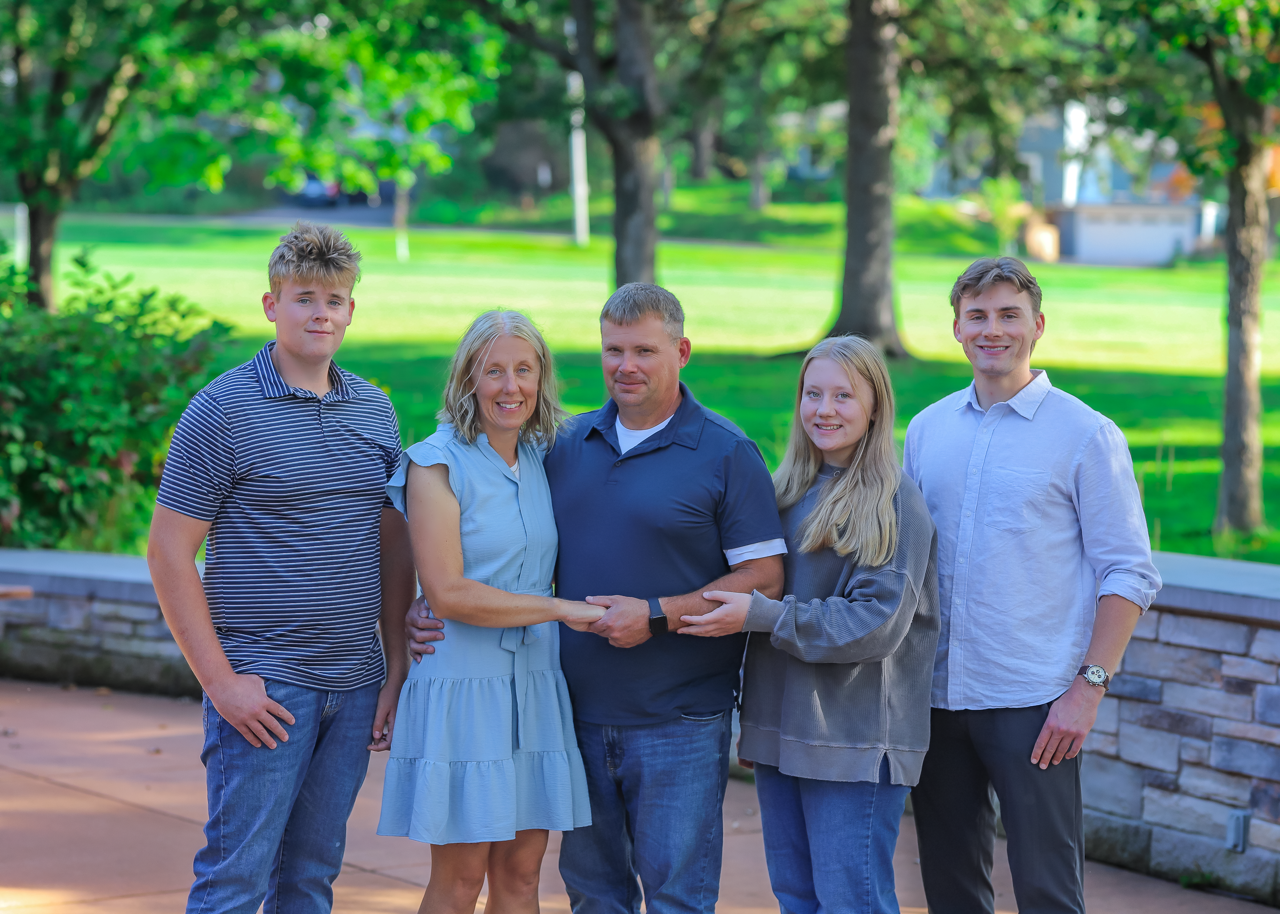 A group of five people stand outdoors on a stone patio with a park in the background, smiling together for a photo.