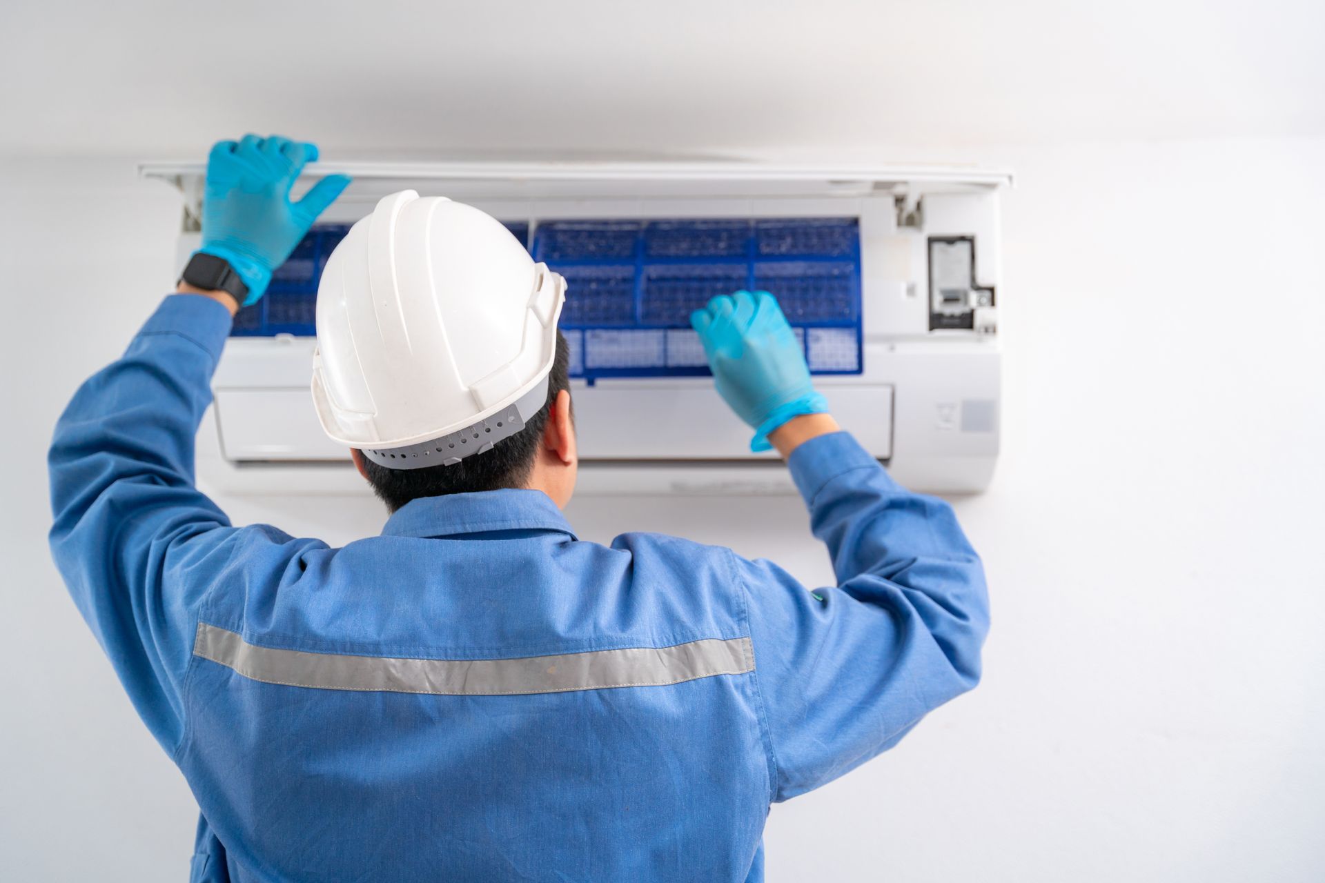 A technician in a blue uniform and white hard hat services a wall-mounted air conditioning unit.