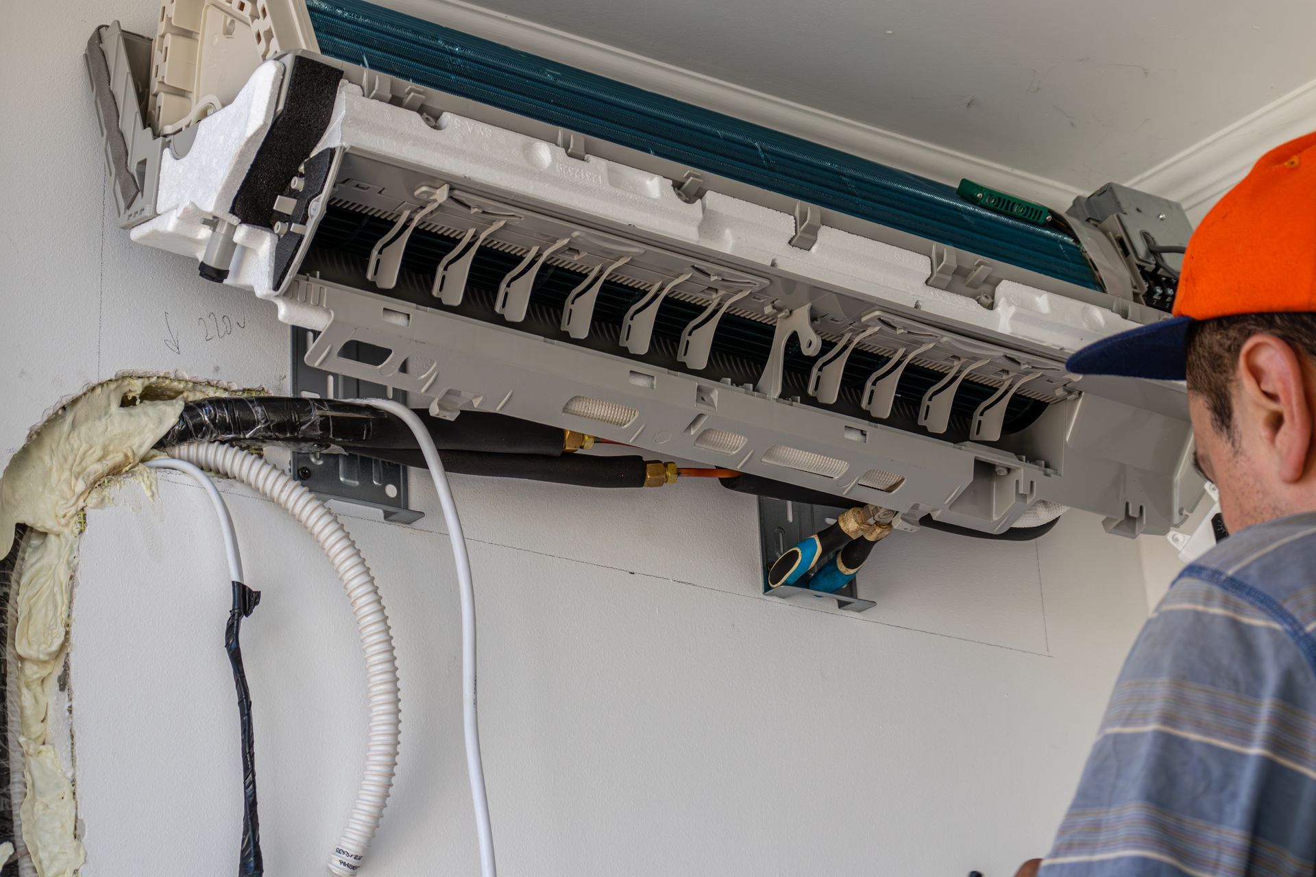 A worker wearing an orange hat installs a wall-mounted air conditioning unit indoors.