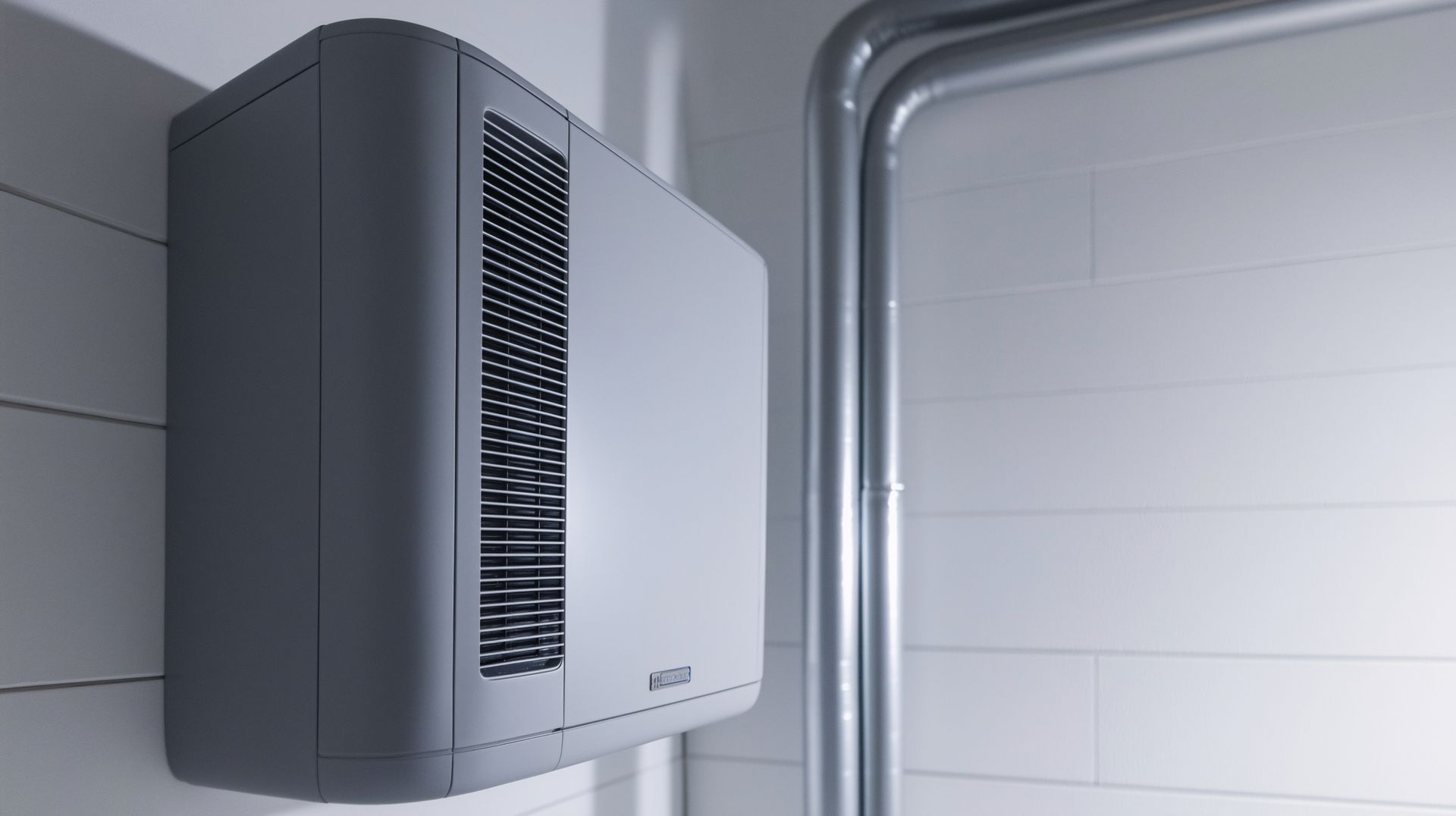 A modern, grey wall-mounted HVAC unit with a side vent installed next to metal pipes on a white tiled wall.