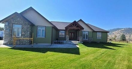 Green house with stone facade, wooden porch, and lawn, against a mountain backdrop.