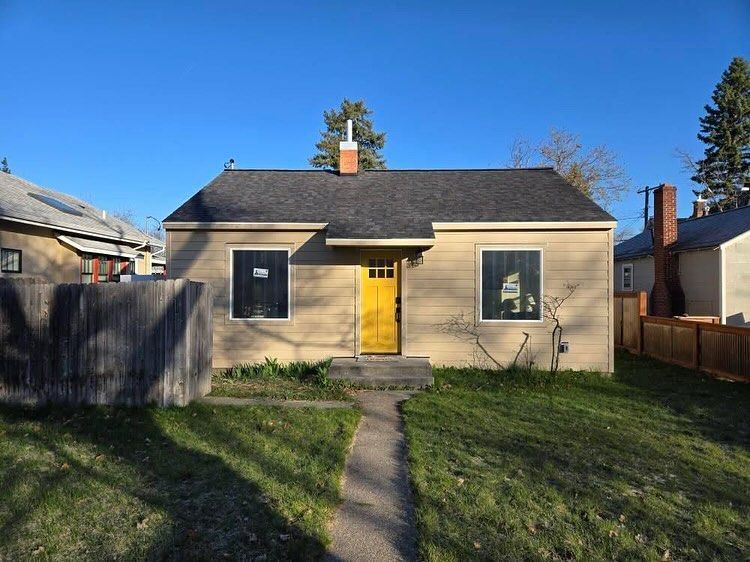 Tan bungalow with a yellow door, black windows, and a chimney on a sunny day.