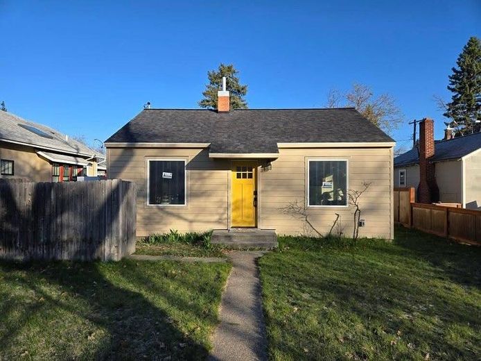 Tan bungalow with a yellow door, black windows, and a chimney on a sunny day.