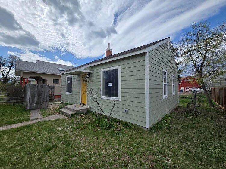 Green house with yellow door, grey roof, two windows covered, cloudy sky.
