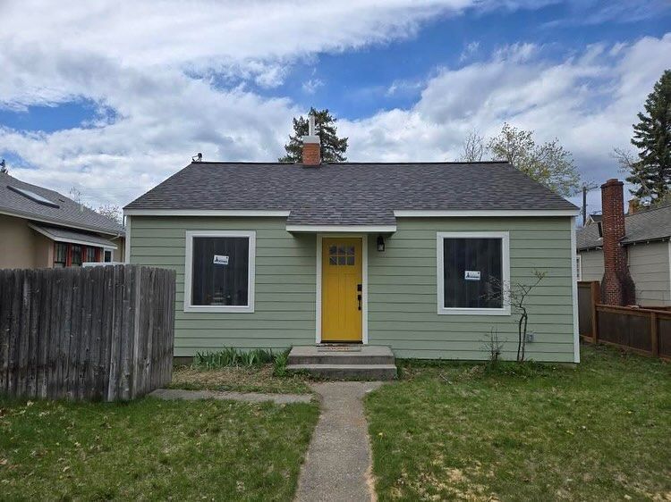 Green house with yellow door and two windows, on a cloudy day. Concrete path leads to steps.