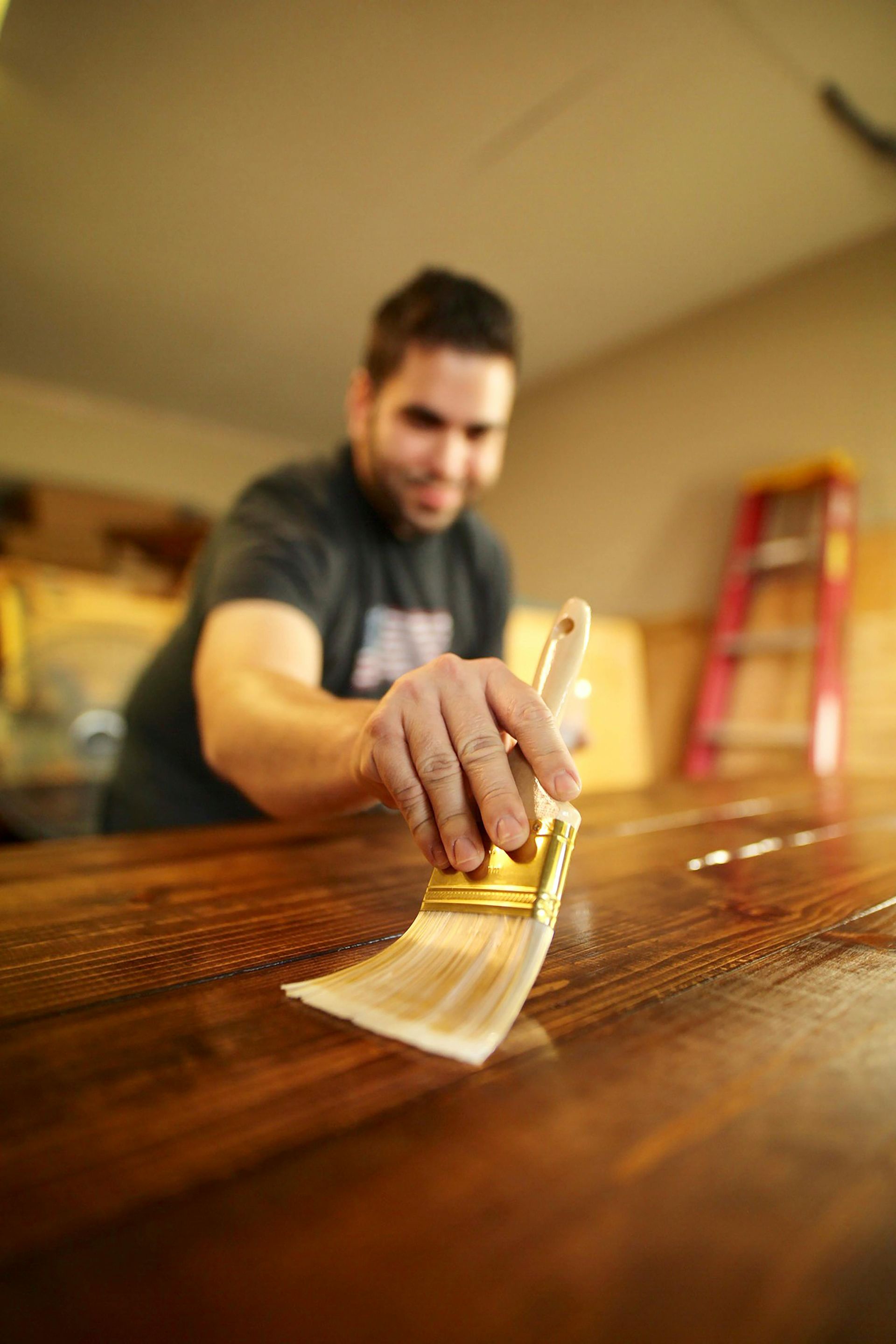 Man applying varnish to a wooden table with a brush. Inside, with a ladder in the background.