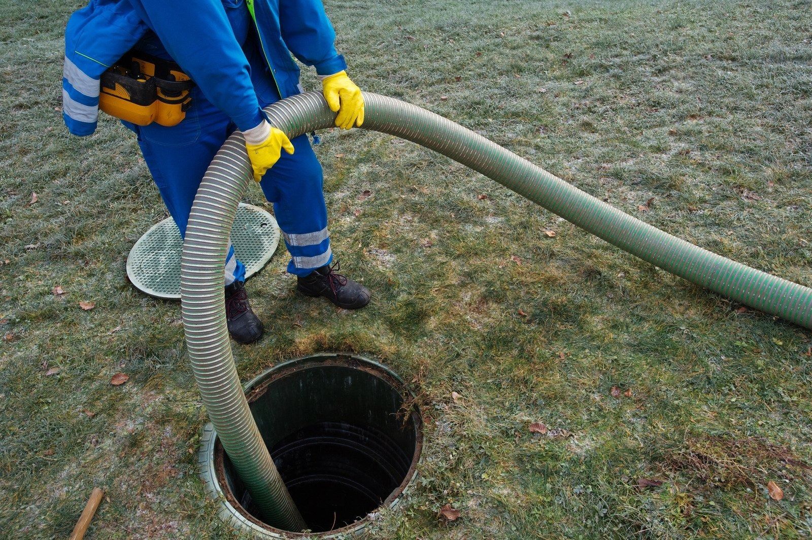 Worker in blue overalls and yellow gloves using a hose at an open manhole on grassy ground