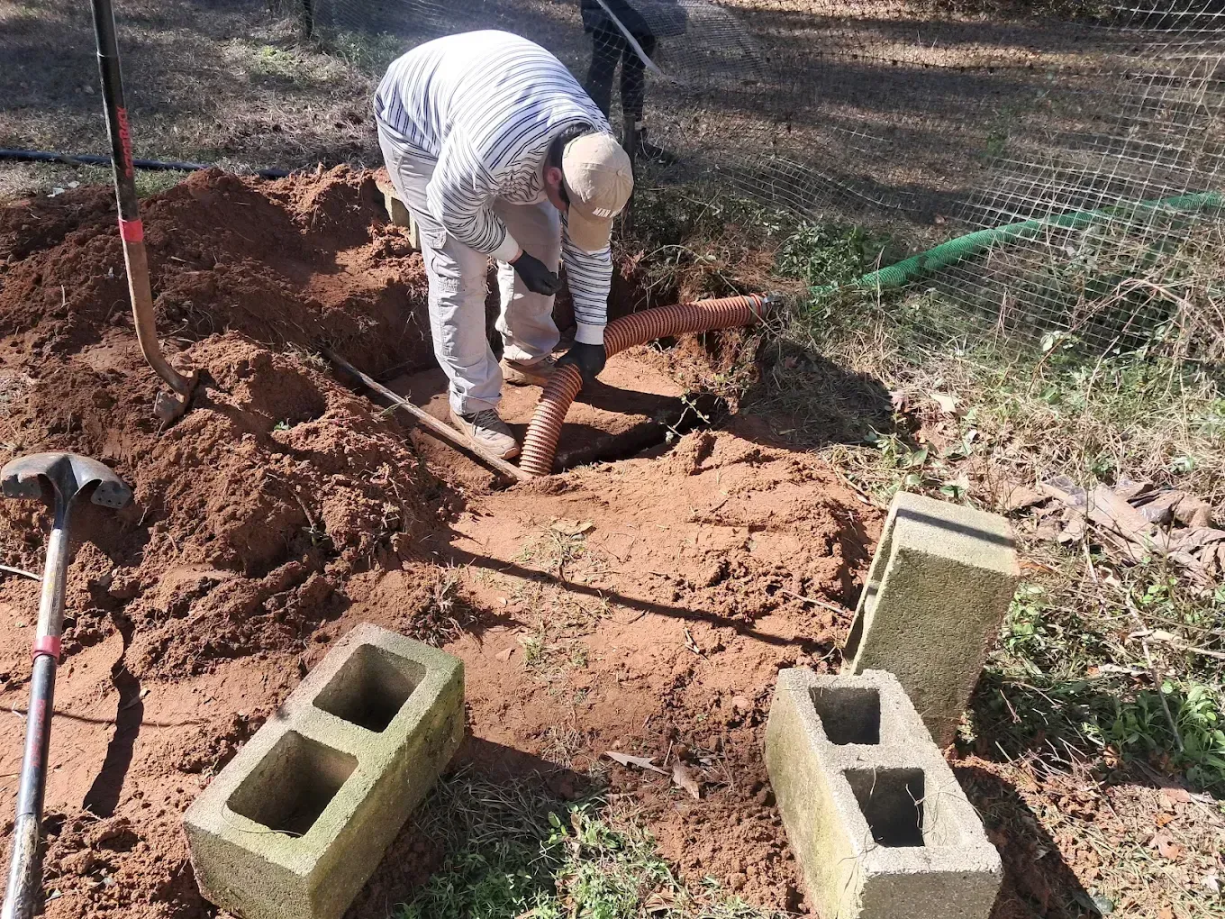 Person digging in a dirt trench beside cinder blocks and tools in a yard.