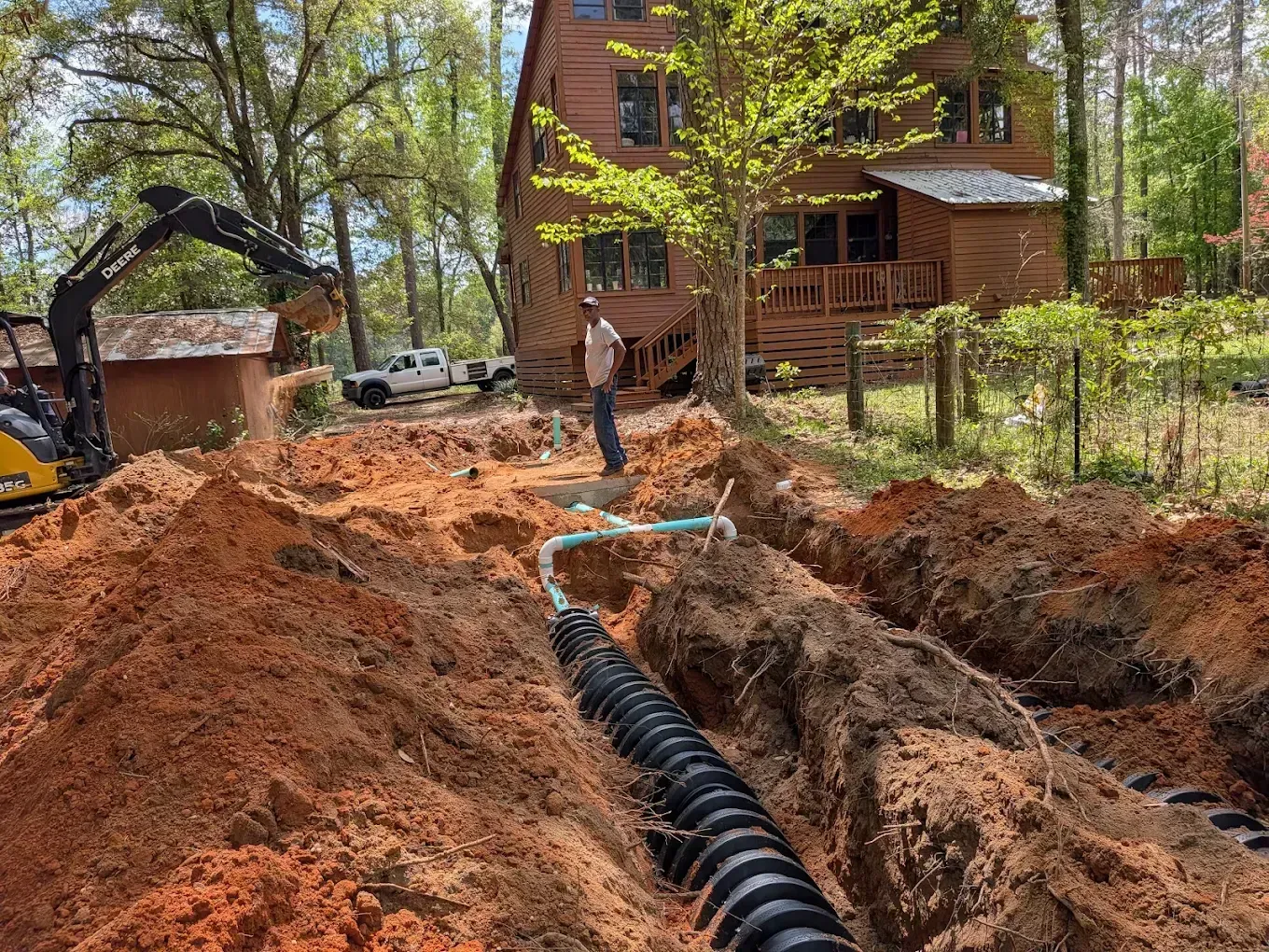 Excavator and worker digging a trench for underground piping beside a house in a wooded yard