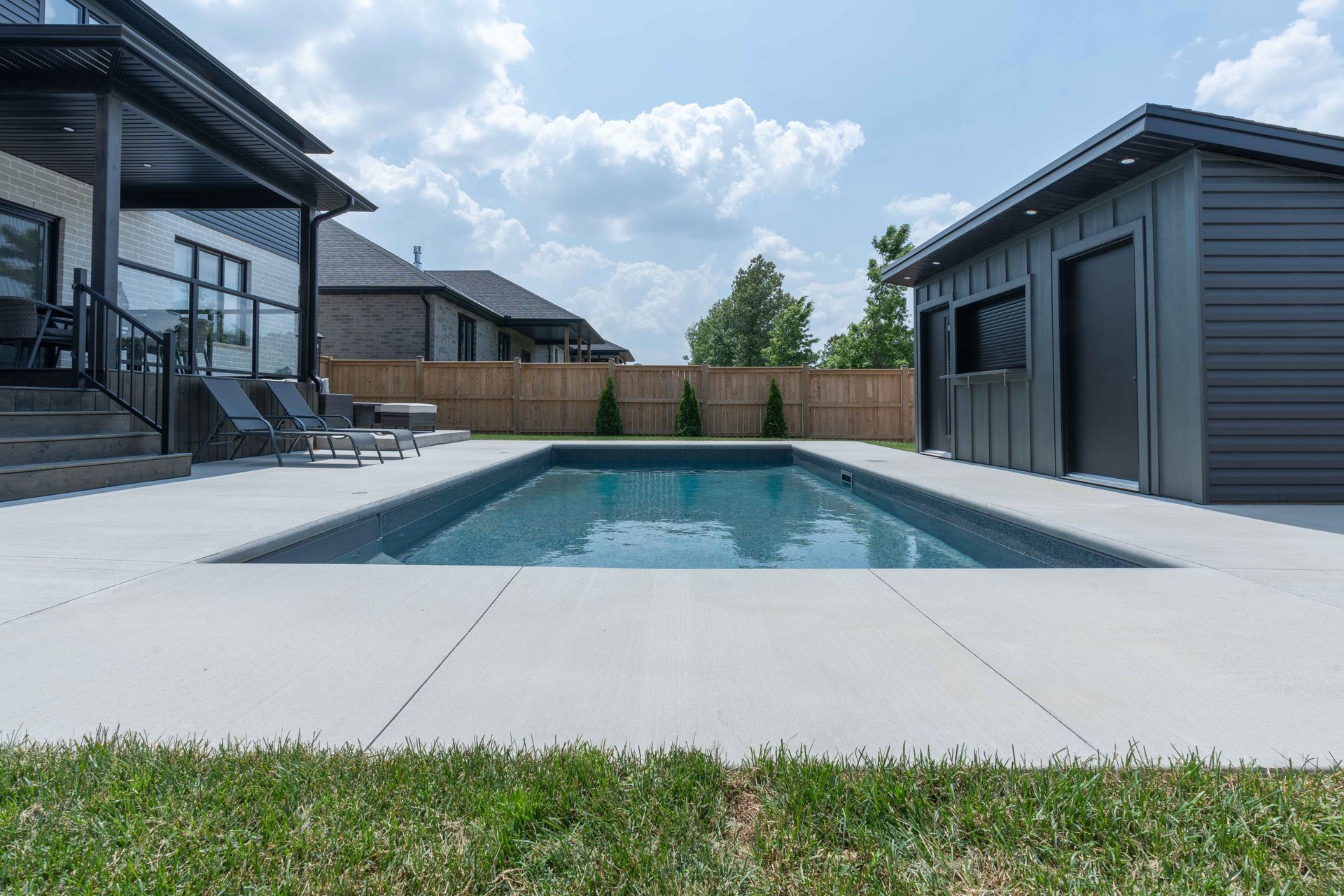Backyard swimming pool between modern gray houses with a wooden fence and grassy foreground