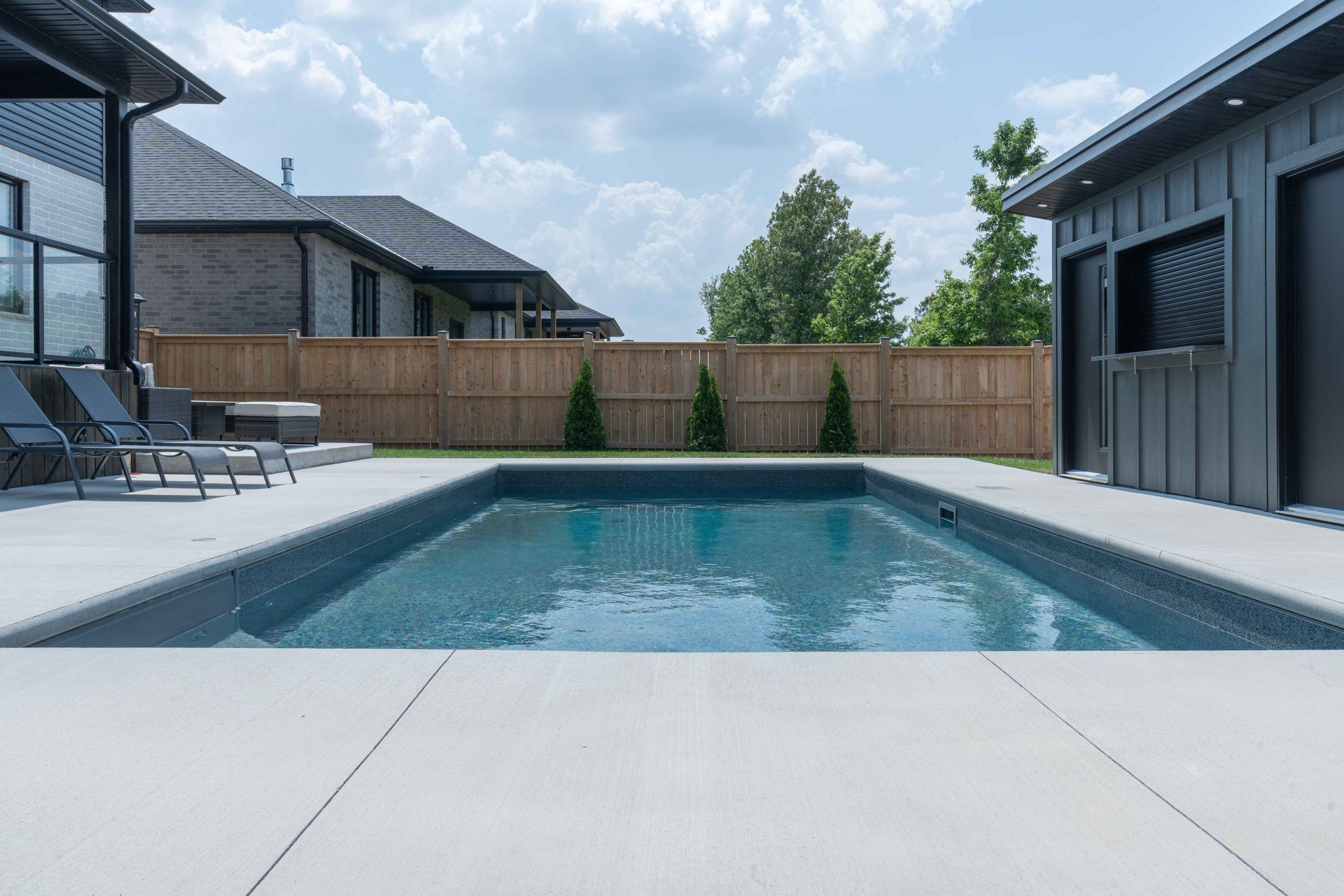 Modern backyard with a rectangular swimming pool, gray patio, lounge chairs, and a wooden privacy fence