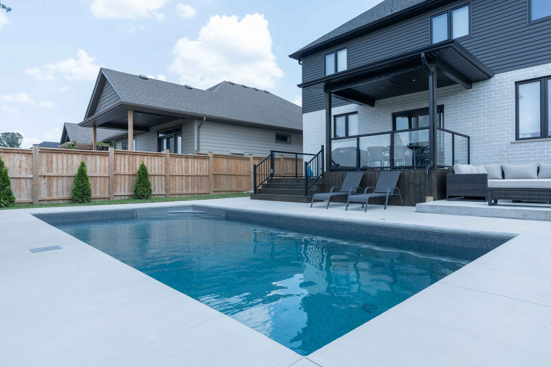 Modern backyard with a rectangular swimming pool beside a white two-story house and patio seating.