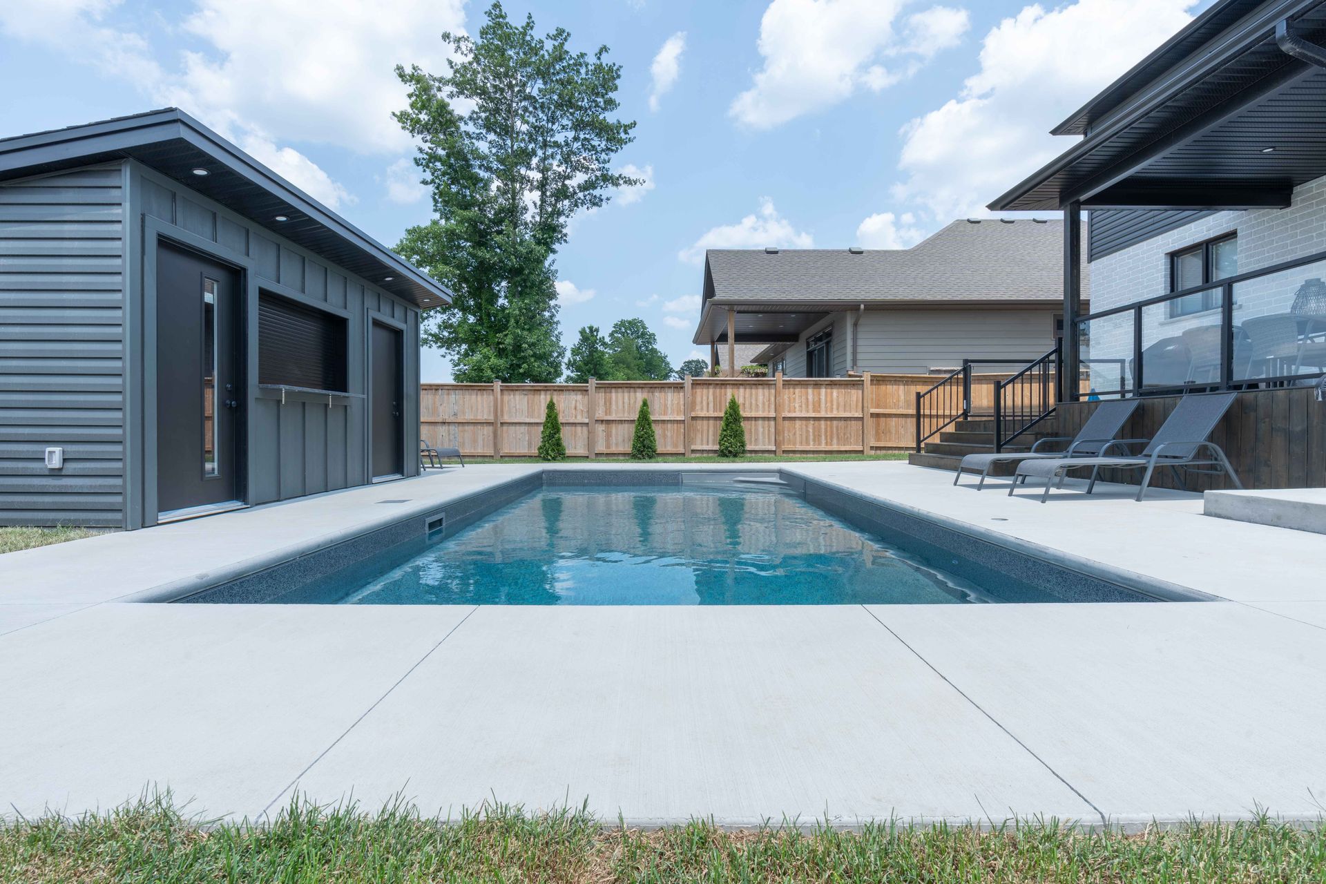 Modern backyard with a rectangular swimming pool, gray pool house, and patio chairs under a blue sky.
