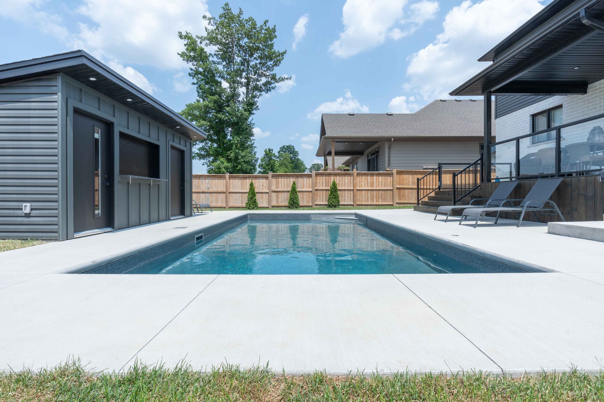 Modern backyard with rectangular swimming pool, gray pool house, wooden fence, and patio seating