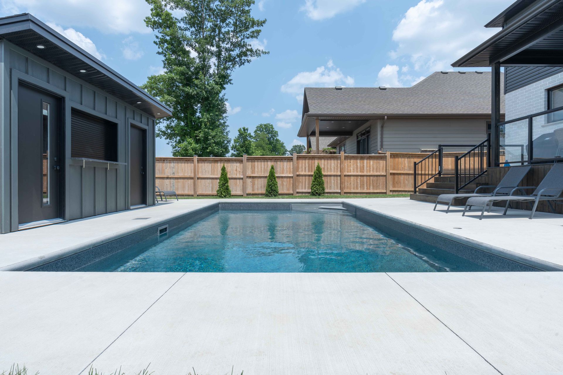Modern backyard pool with lounge chairs, wooden fence, and contemporary house under a blue sky