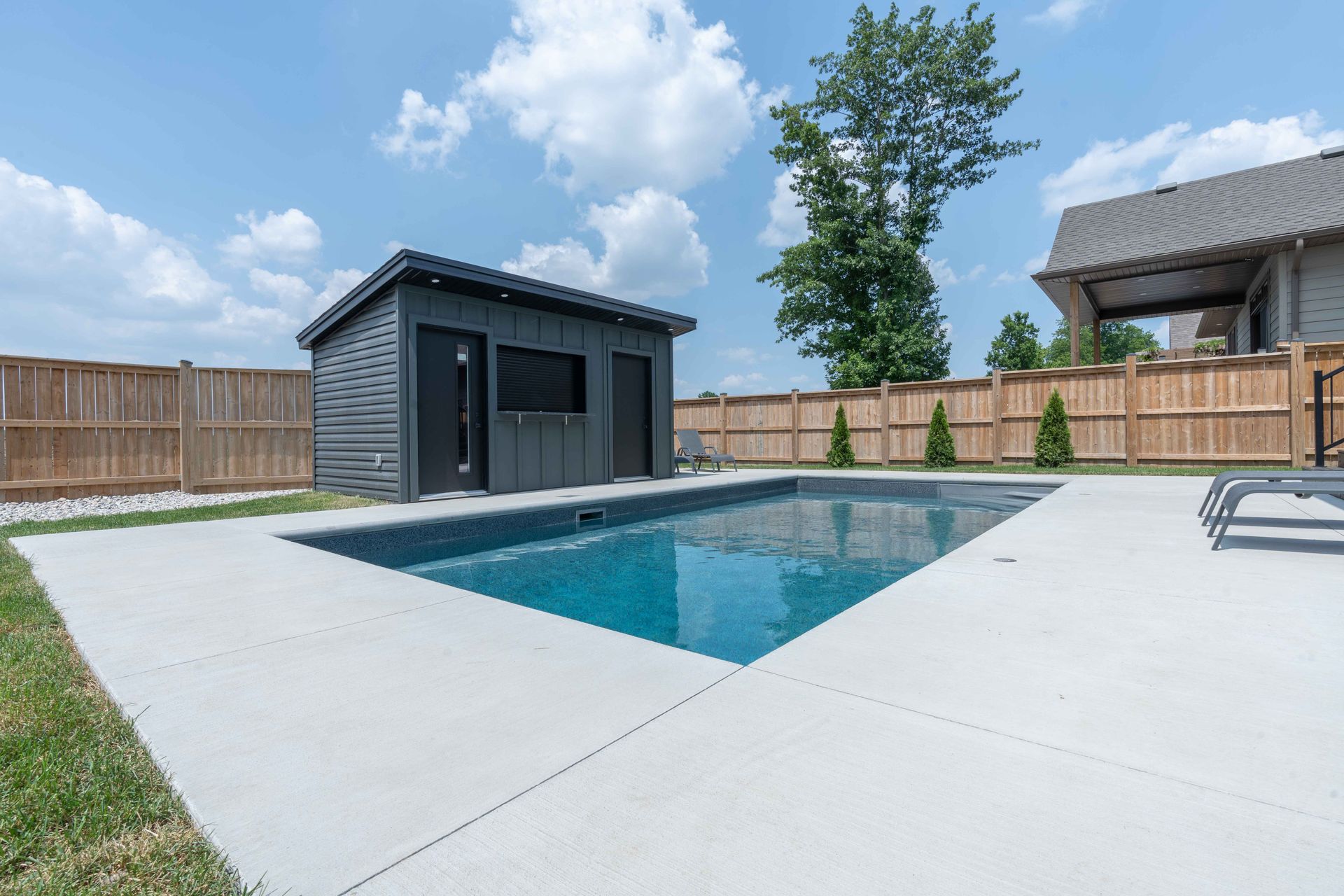Modern backyard with a rectangular pool, concrete patio, and a small gray pool house under a blue sky