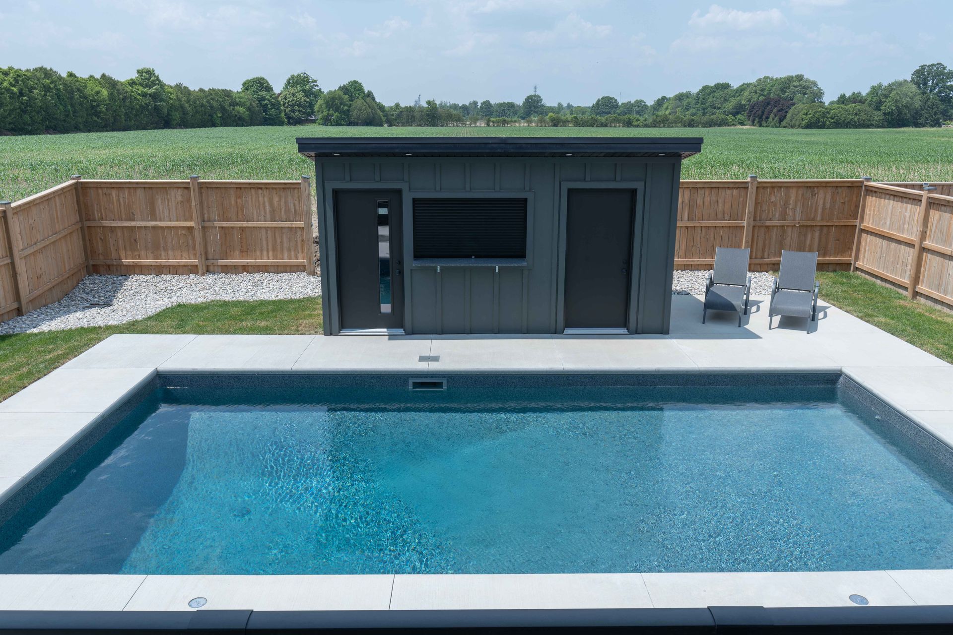 Backyard pool with a dark pool house, lounge chairs, and fenced lawn beyond