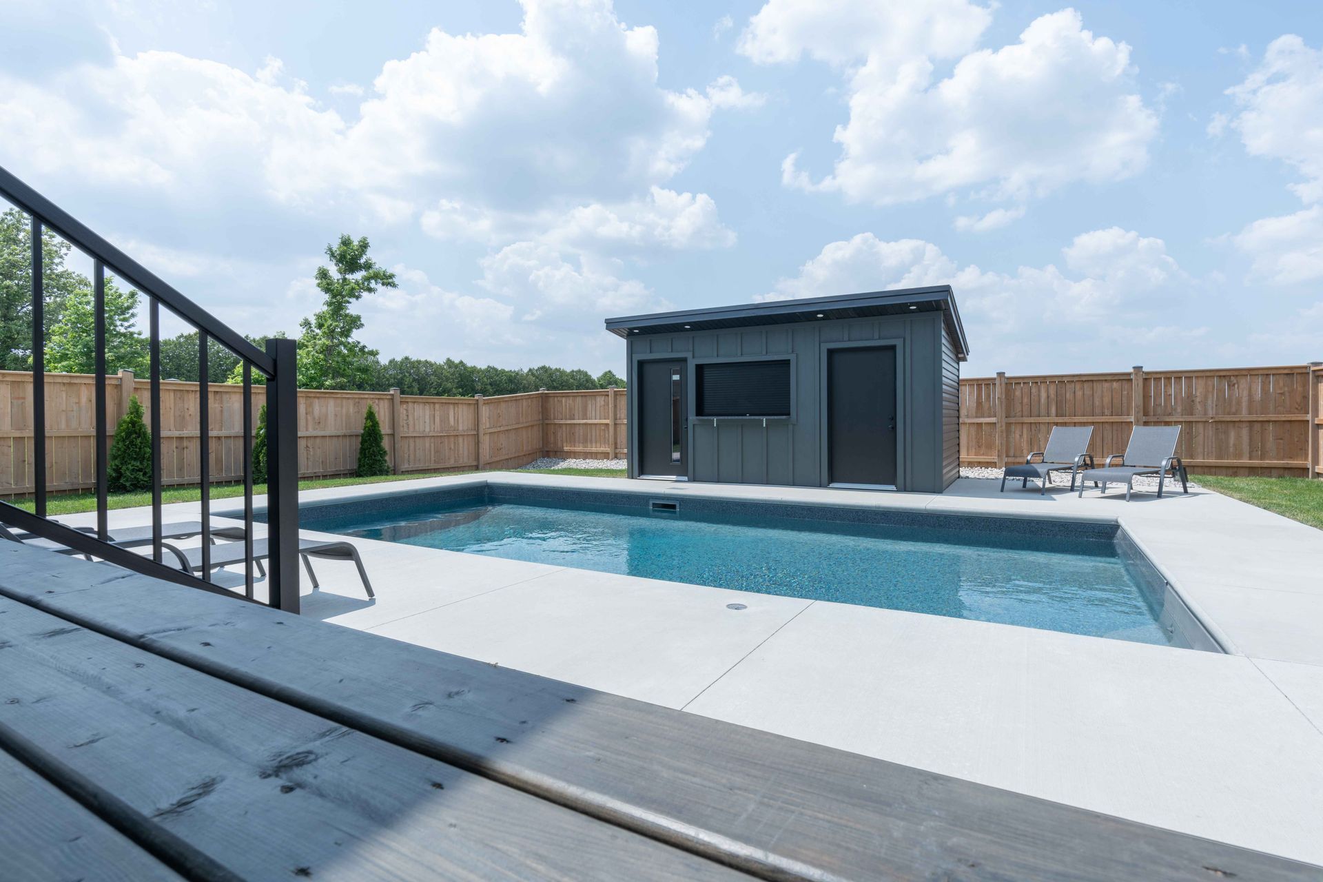 Backyard pool with white deck, dark shed, and fenced yard under a cloudy sky