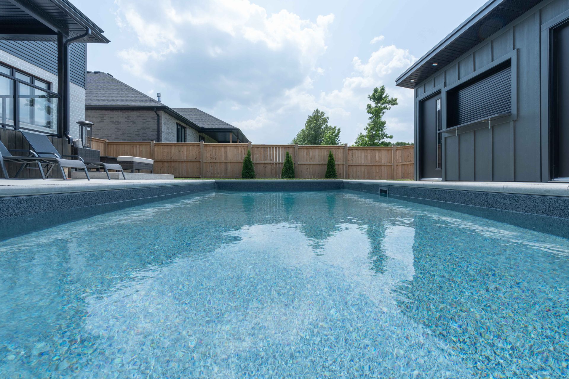 Backyard swimming pool with patio chairs and modern houses under a cloudy sky