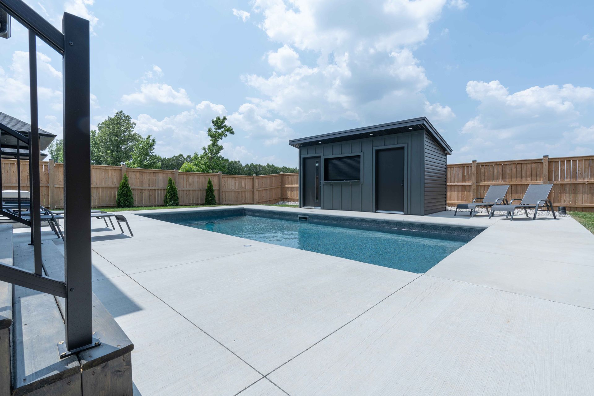 Modern backyard with rectangular swimming pool, concrete patio, lounge chairs, and a small pool house under a blue sky