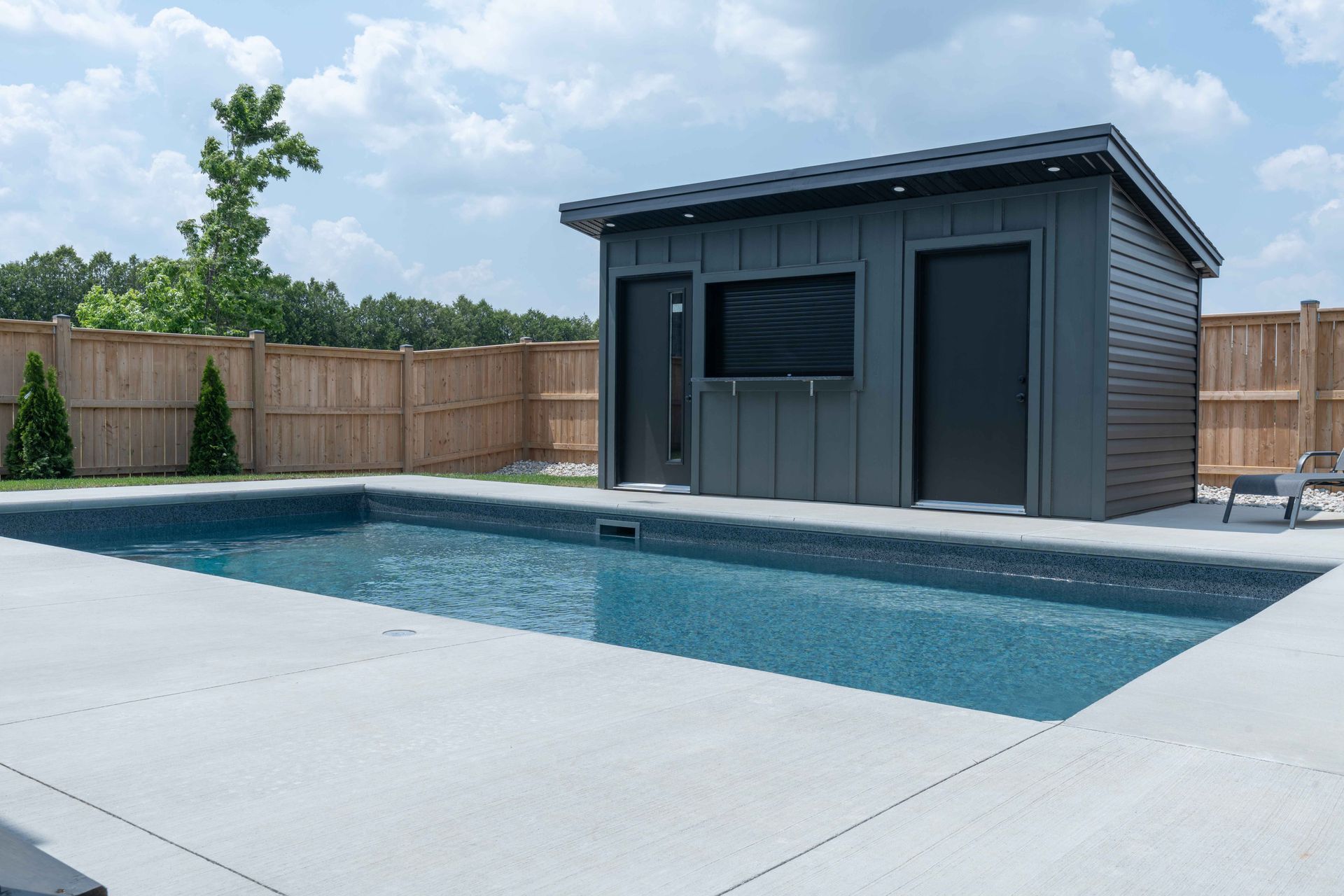 Modern backyard pool beside a gray pool house and concrete patio, with wooden privacy fencing.