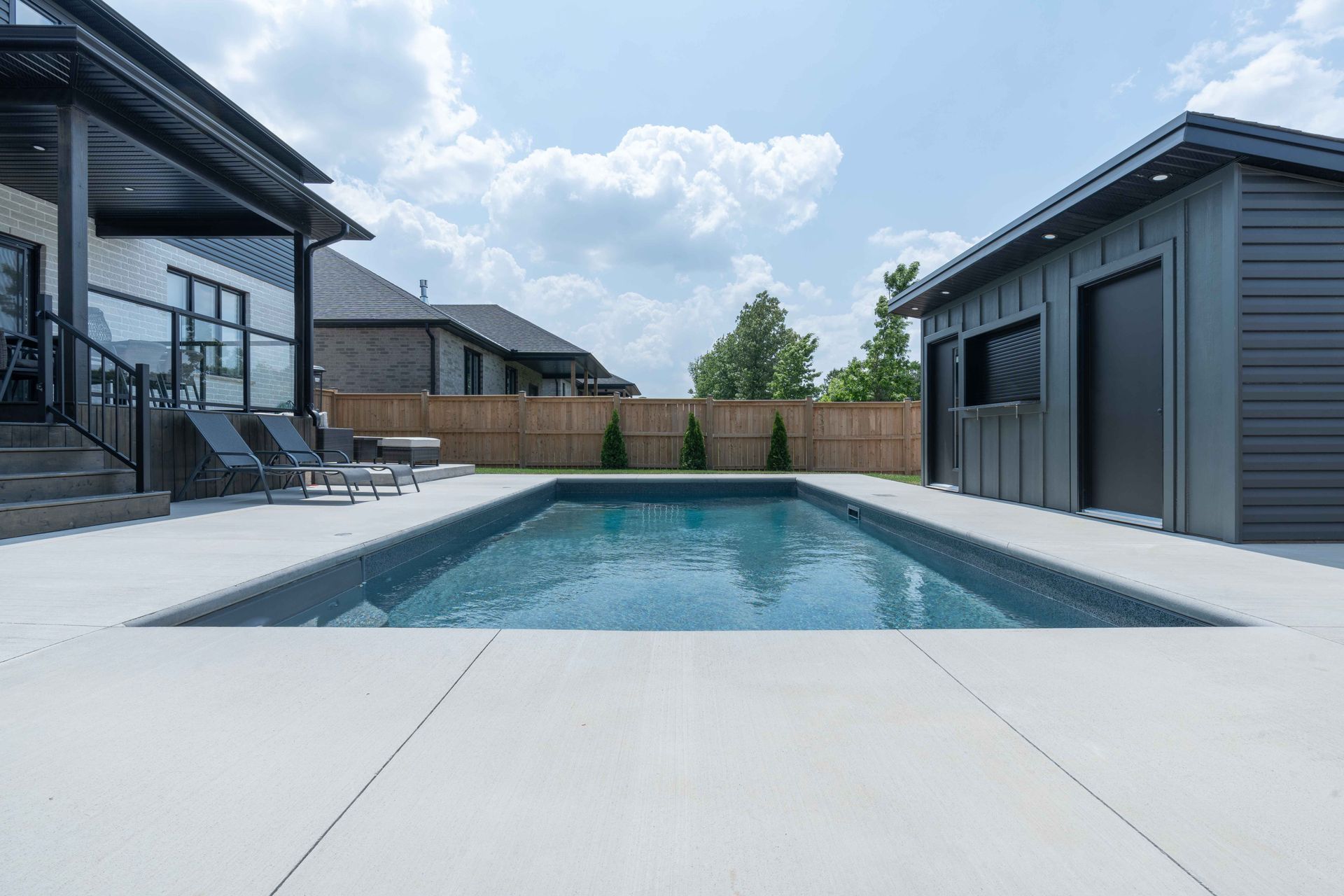 Modern backyard with a rectangular swimming pool, patio, and two gray buildings under a partly cloudy sky
