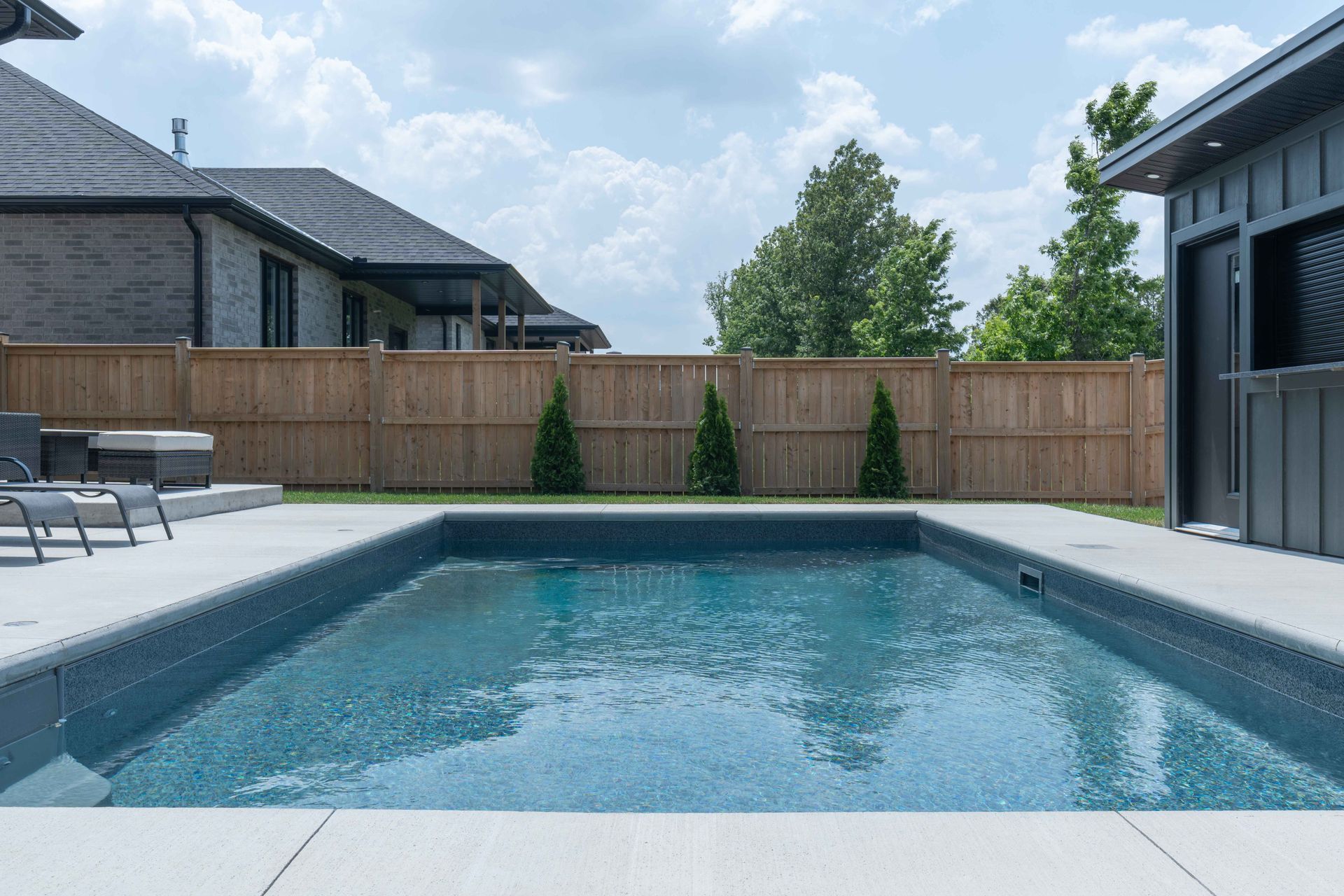 Backyard swimming pool with patio chairs, wooden fence, and house exteriors under a cloudy sky