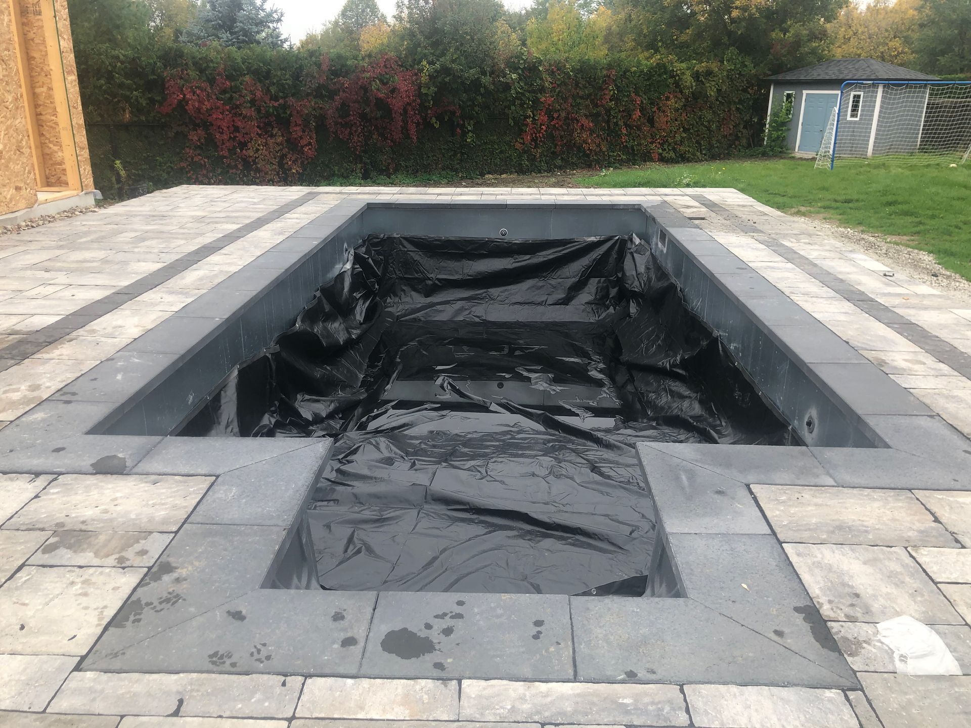 An in-ground swimming pool under construction, lined with black plastic, surrounded by grey stone pavers in a backyard.