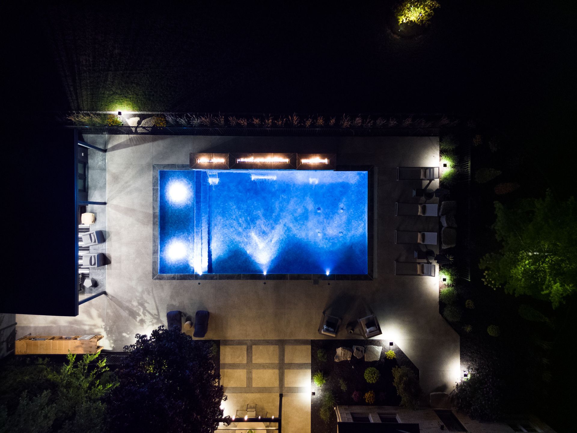 Aerial view of a lit blue swimming pool at night, surrounded by patio and trees.
