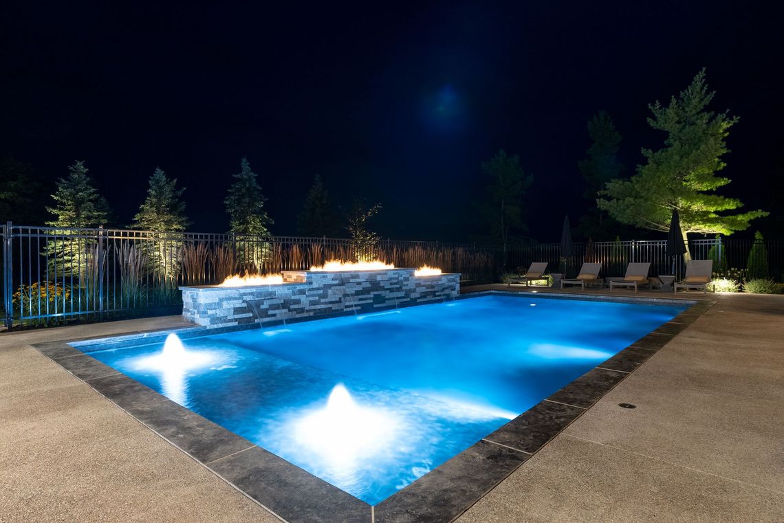 Blue-lit outdoor swimming pool at night with stone deck, lounge chairs, and trees in the background