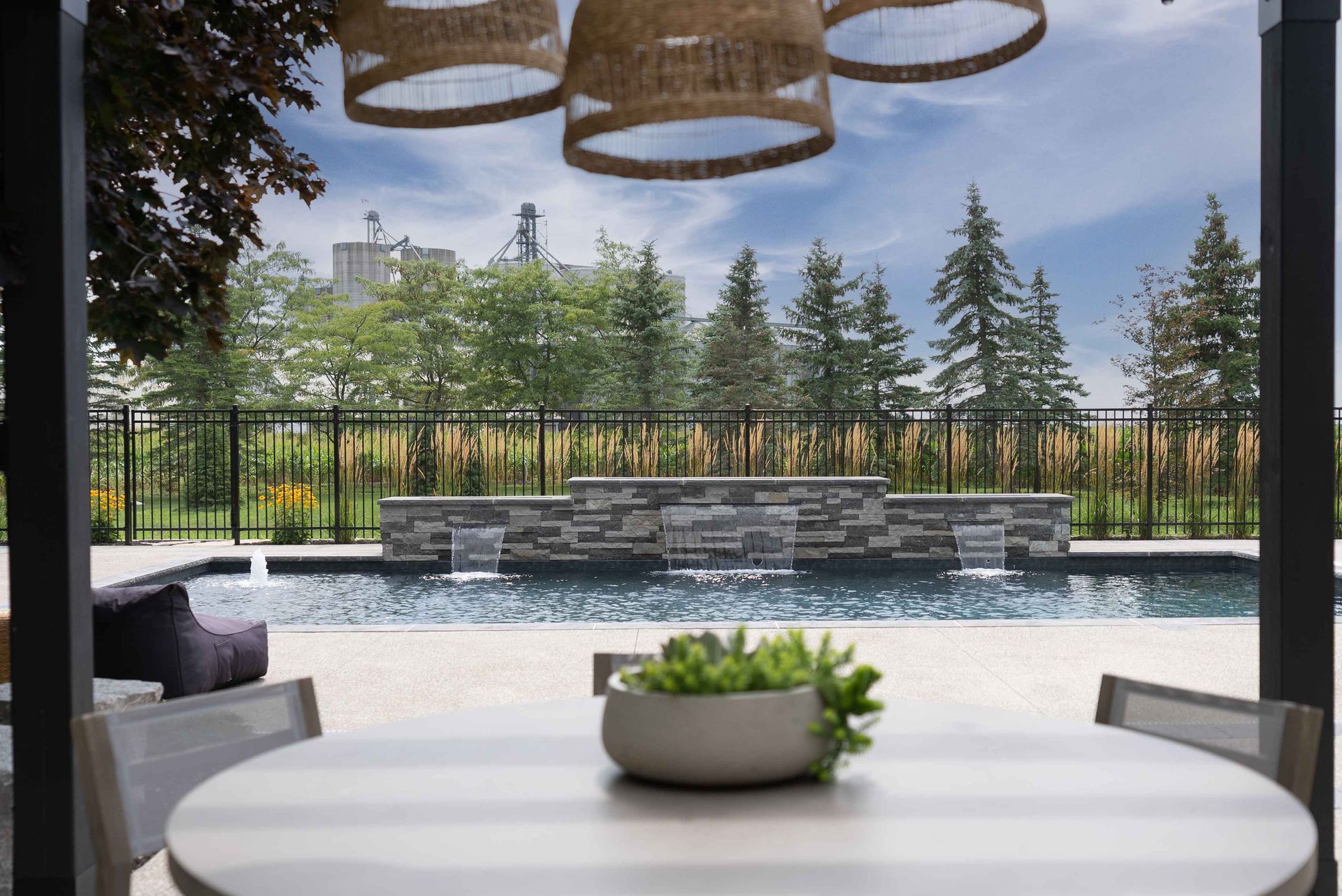 Patio dining table overlooking a pool, stone fountain, and trees under woven hanging lights.