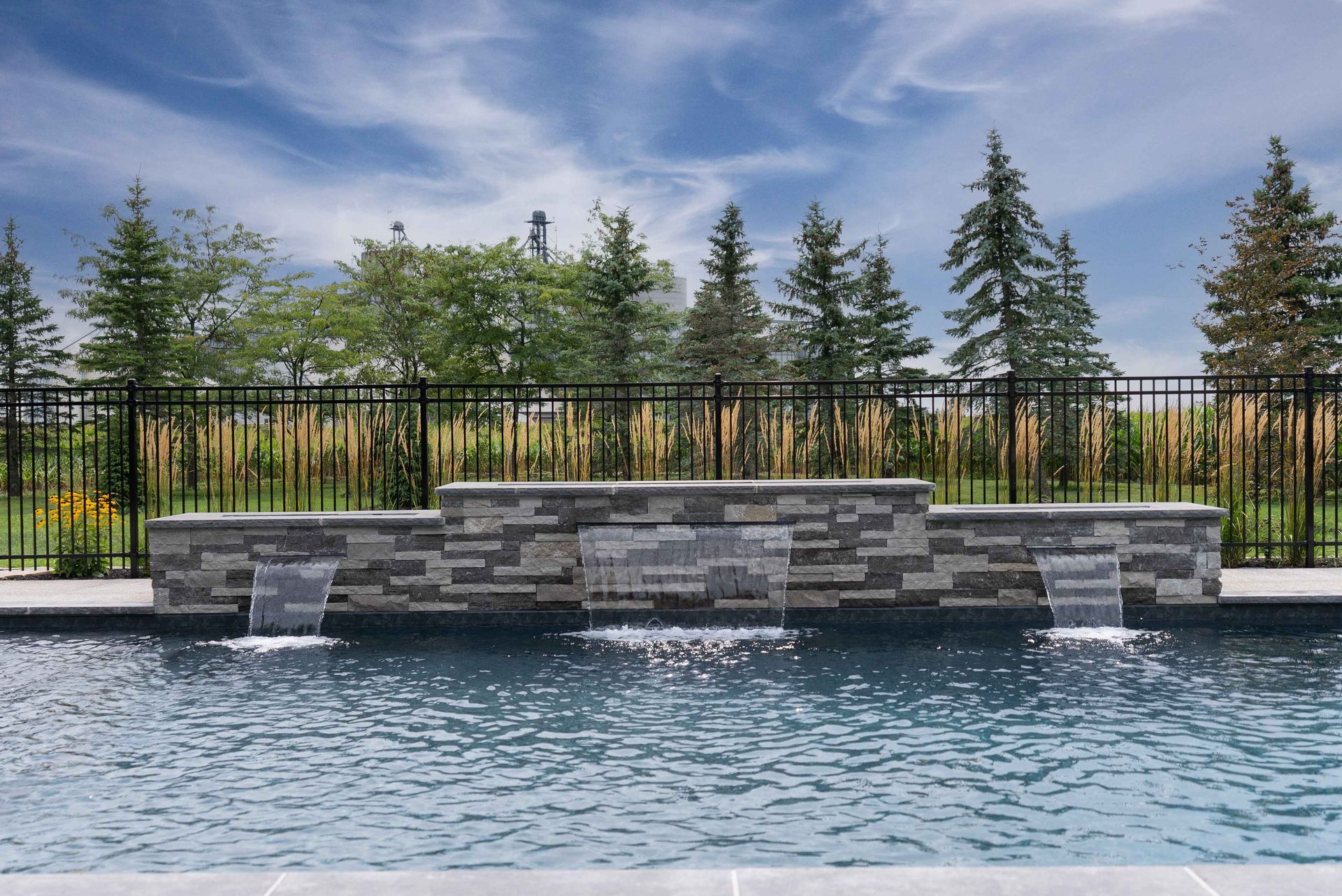 Stone pool fountain with rippling water, black fence, and evergreen trees under a cloudy sky
