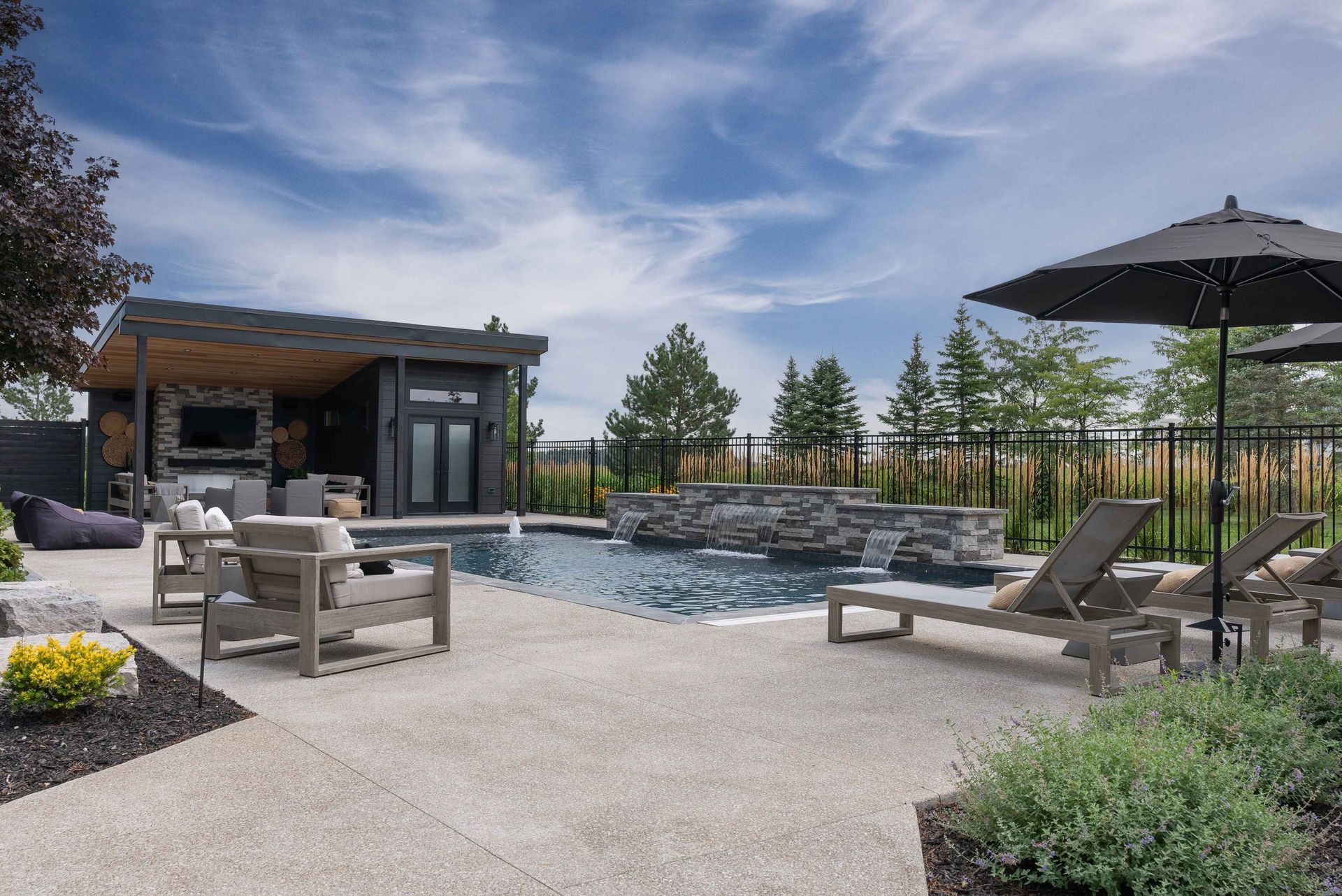 Modern backyard patio with pool, lounge chairs, fire pit, and black umbrella under a cloudy sky