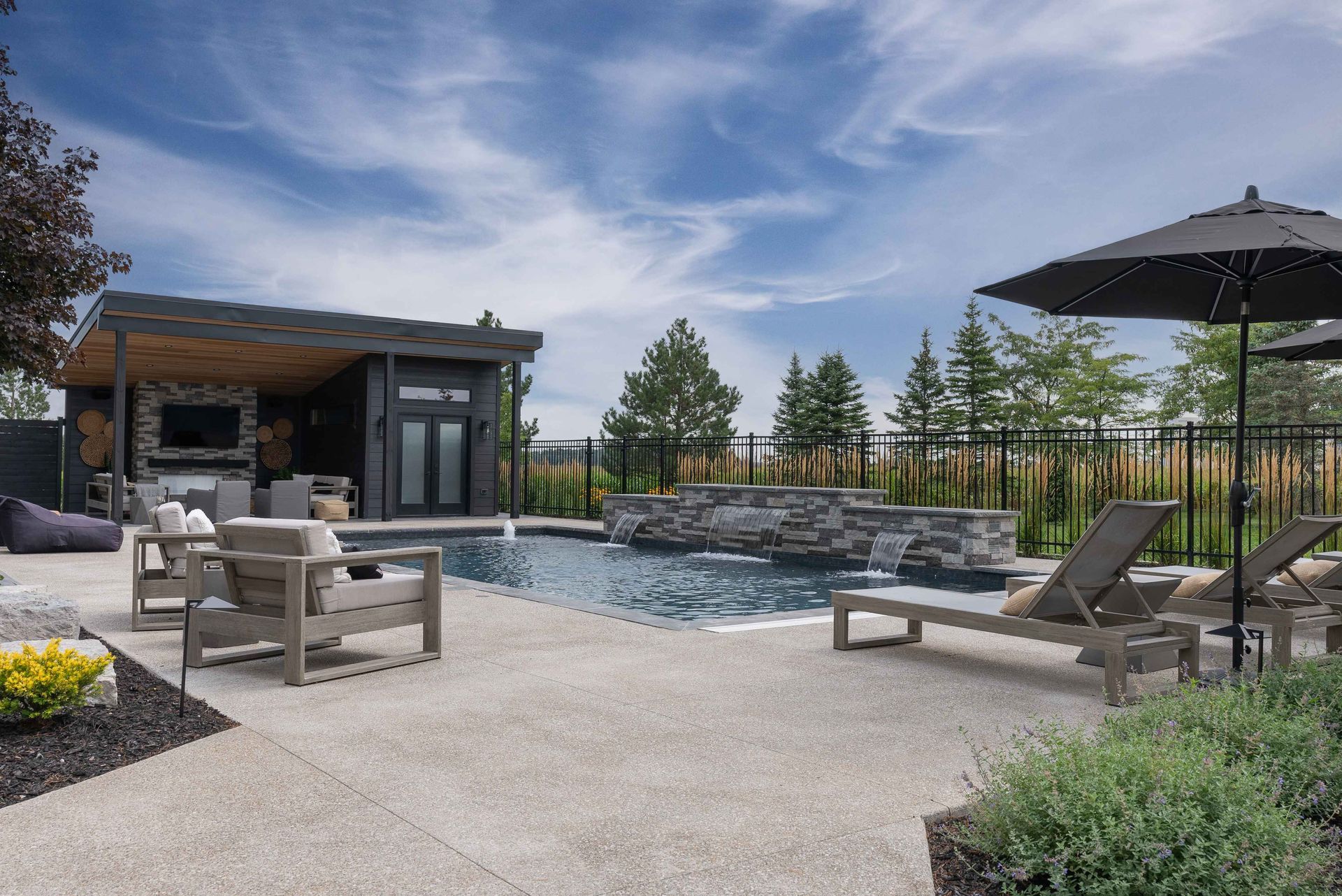 Modern patio with lounge chairs, fire pit, umbrella, and pool beside a small building under a blue sky