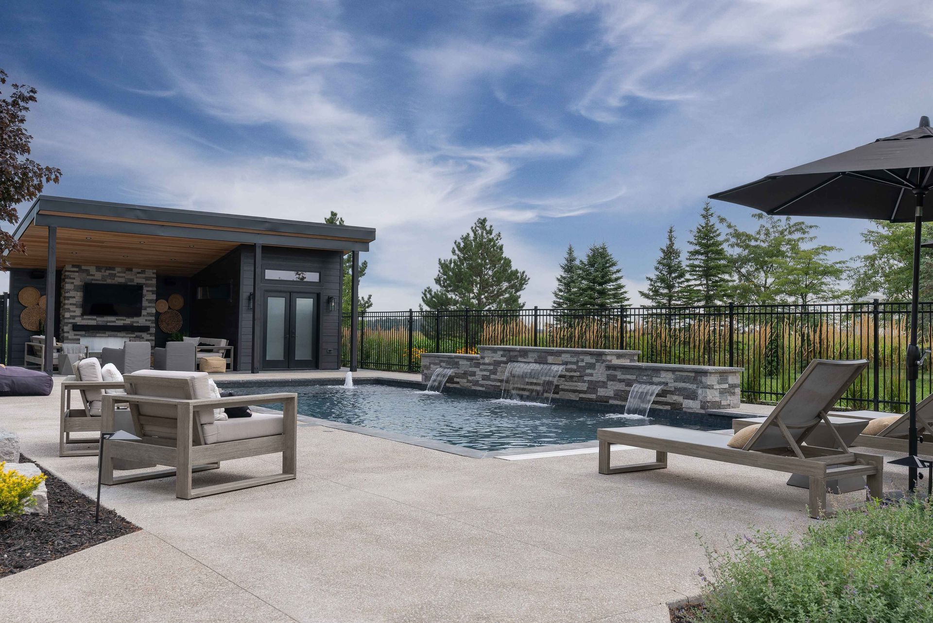 Modern backyard patio with pool, lounge chairs, fire pit, and a small cabin under a blue sky