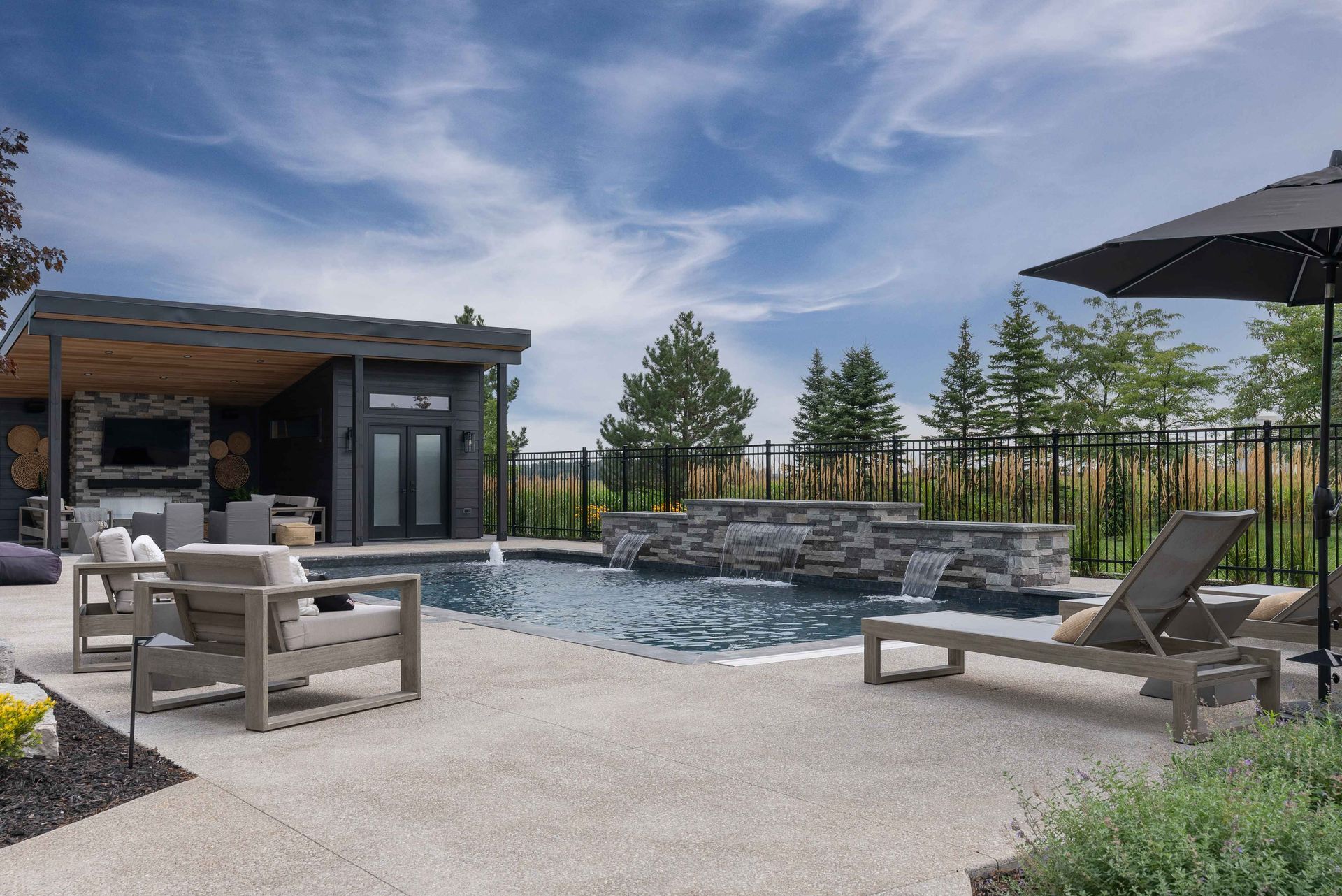 Modern backyard patio with a pool, fire pit, lounge chairs, and a covered seating area under a blue sky