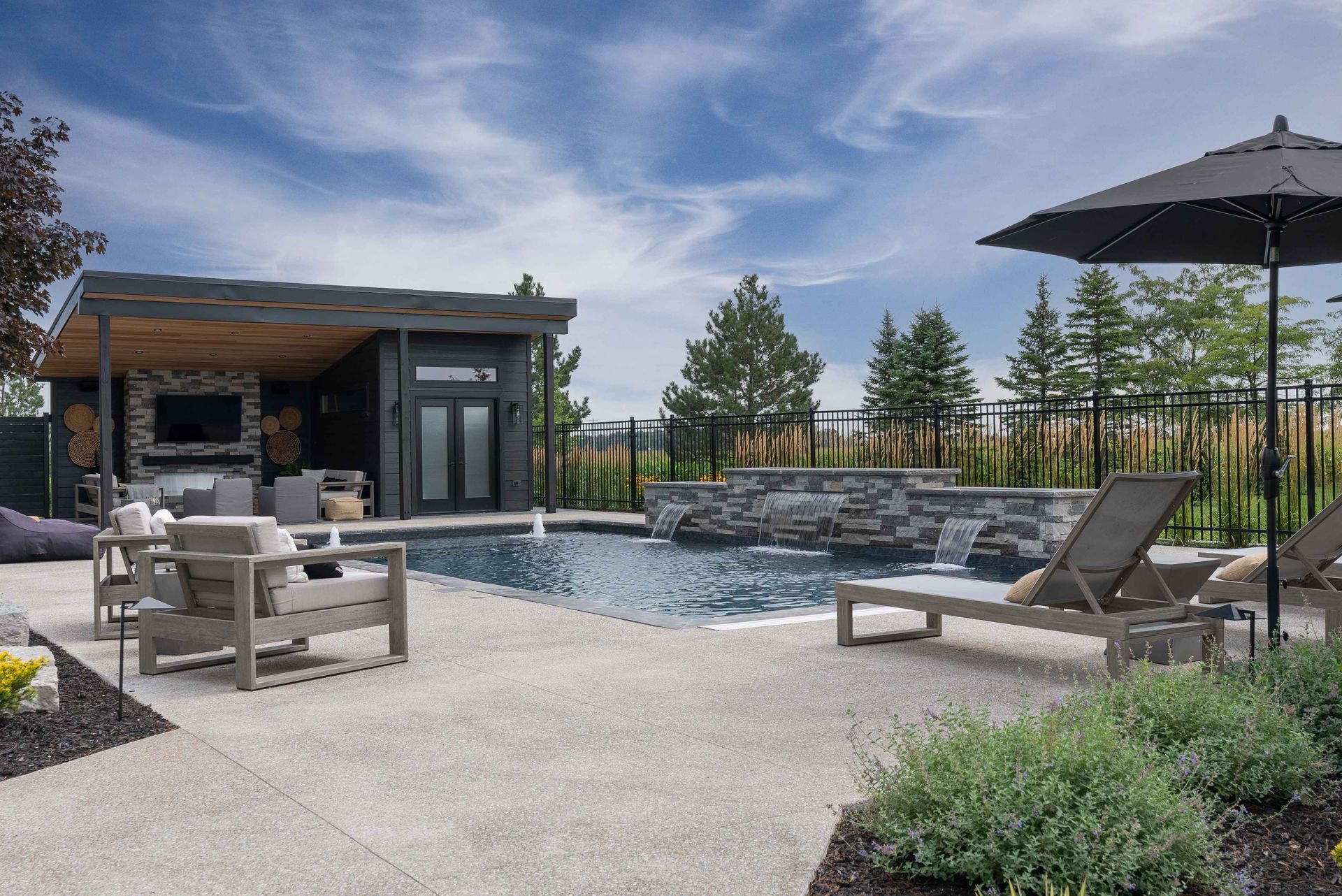 Modern backyard patio with a pool, lounge chairs, fire pit, and umbrella under a blue sky