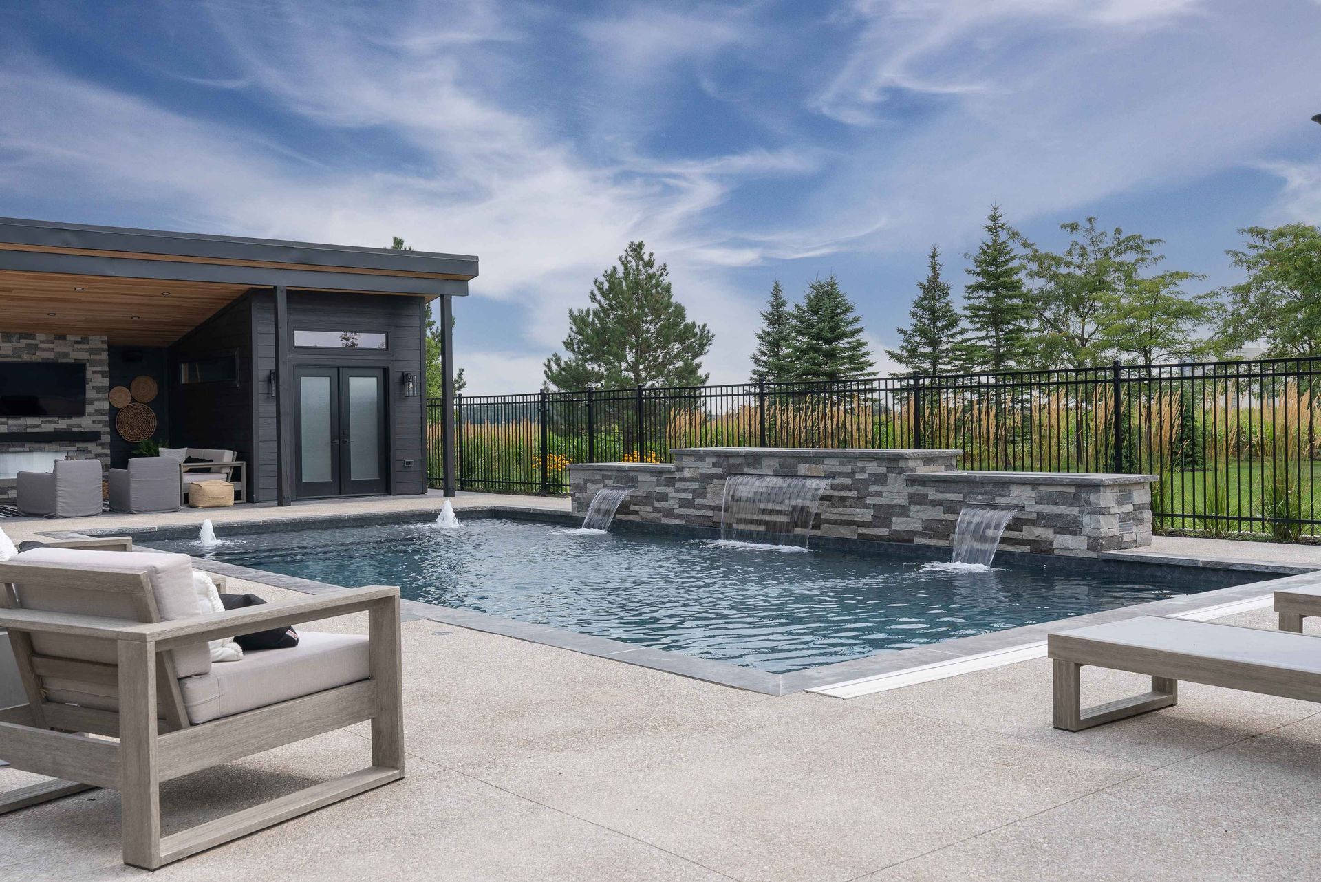Modern backyard pool with stone waterfall, lounge chairs, and pergola under a blue sky
