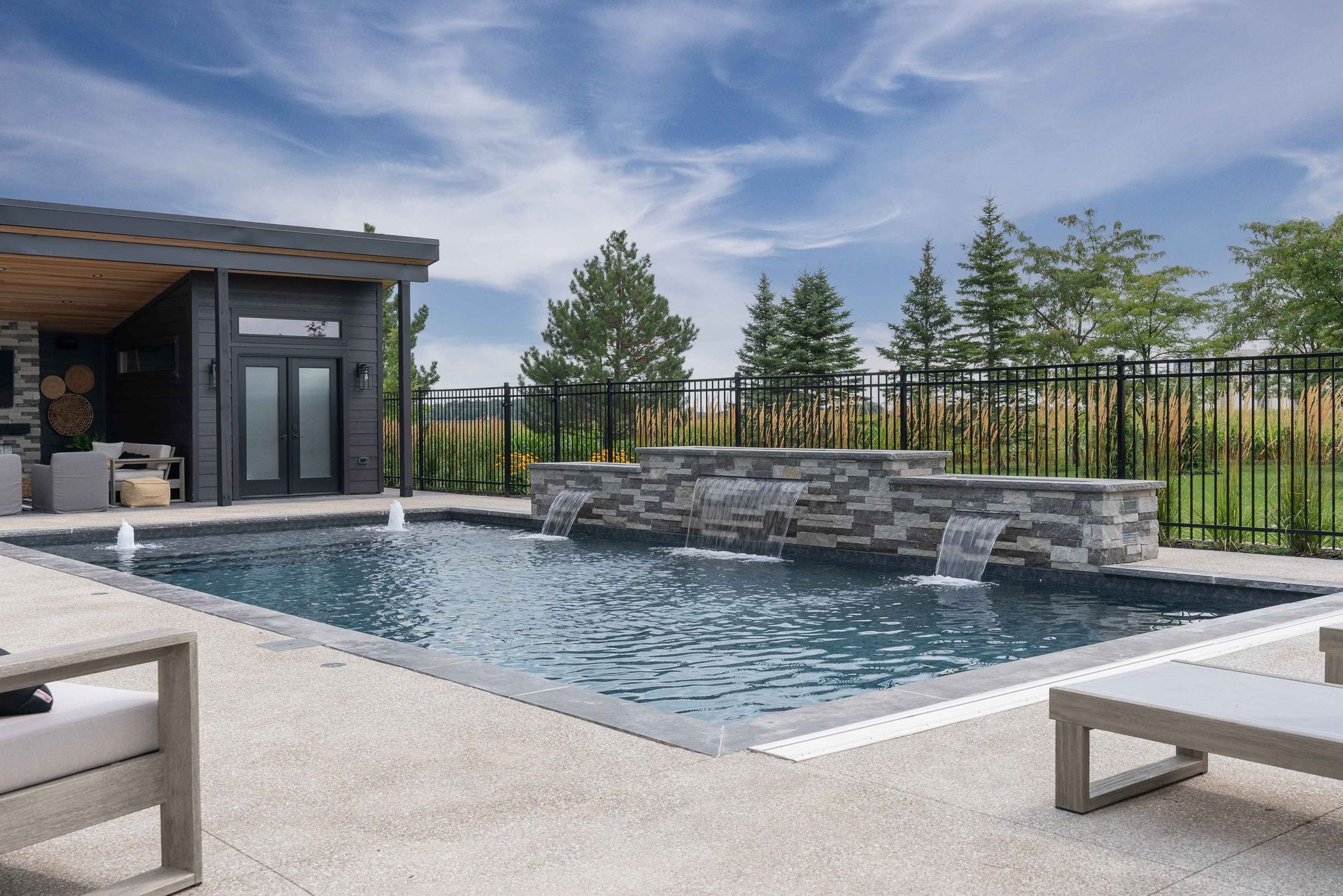 Modern backyard pool with stone waterfall, patio seating, and fenced landscaping under a cloudy sky